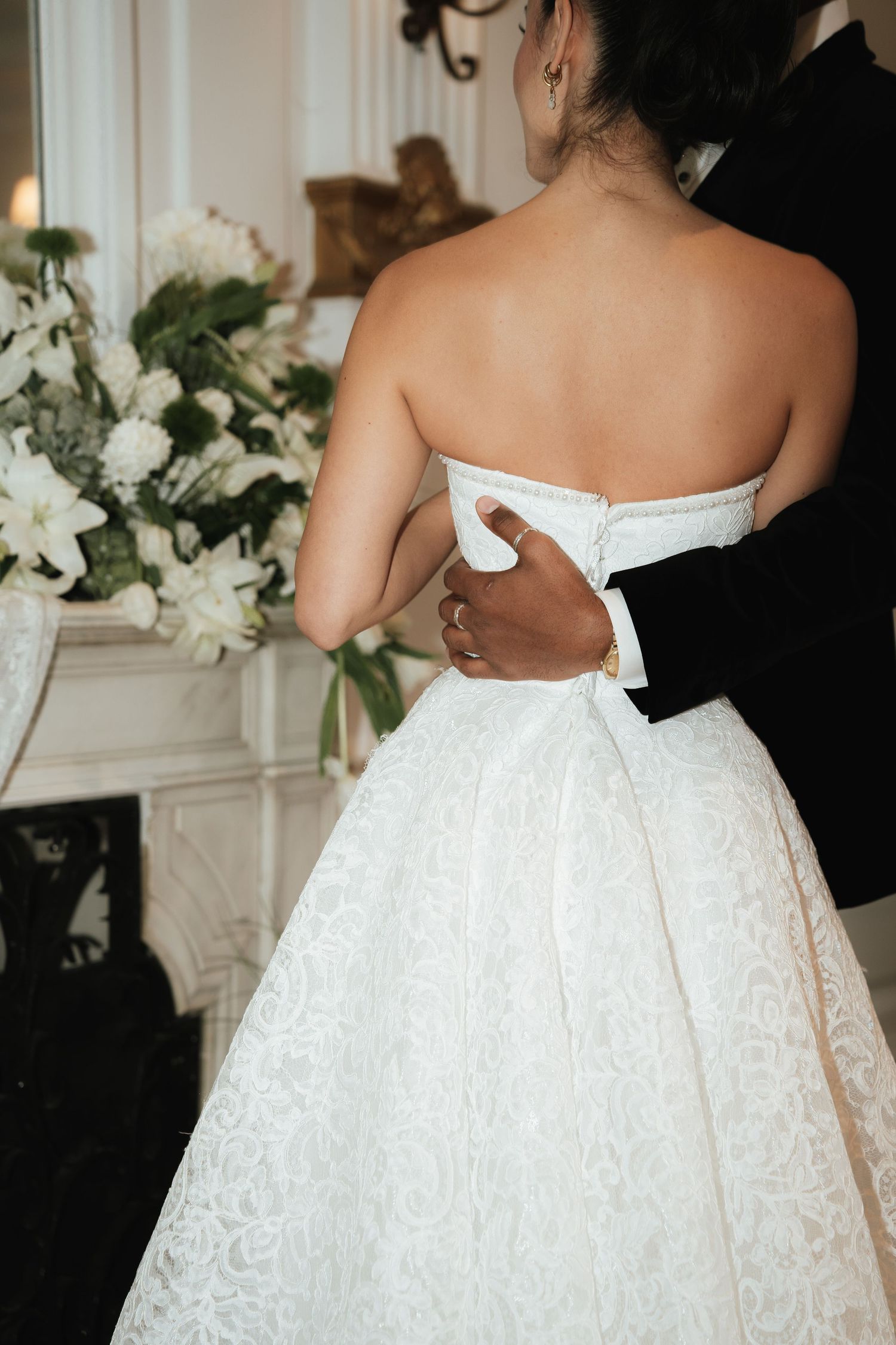 Bride in strapless white lace wedding gown embraced by groom near floral arrangement.