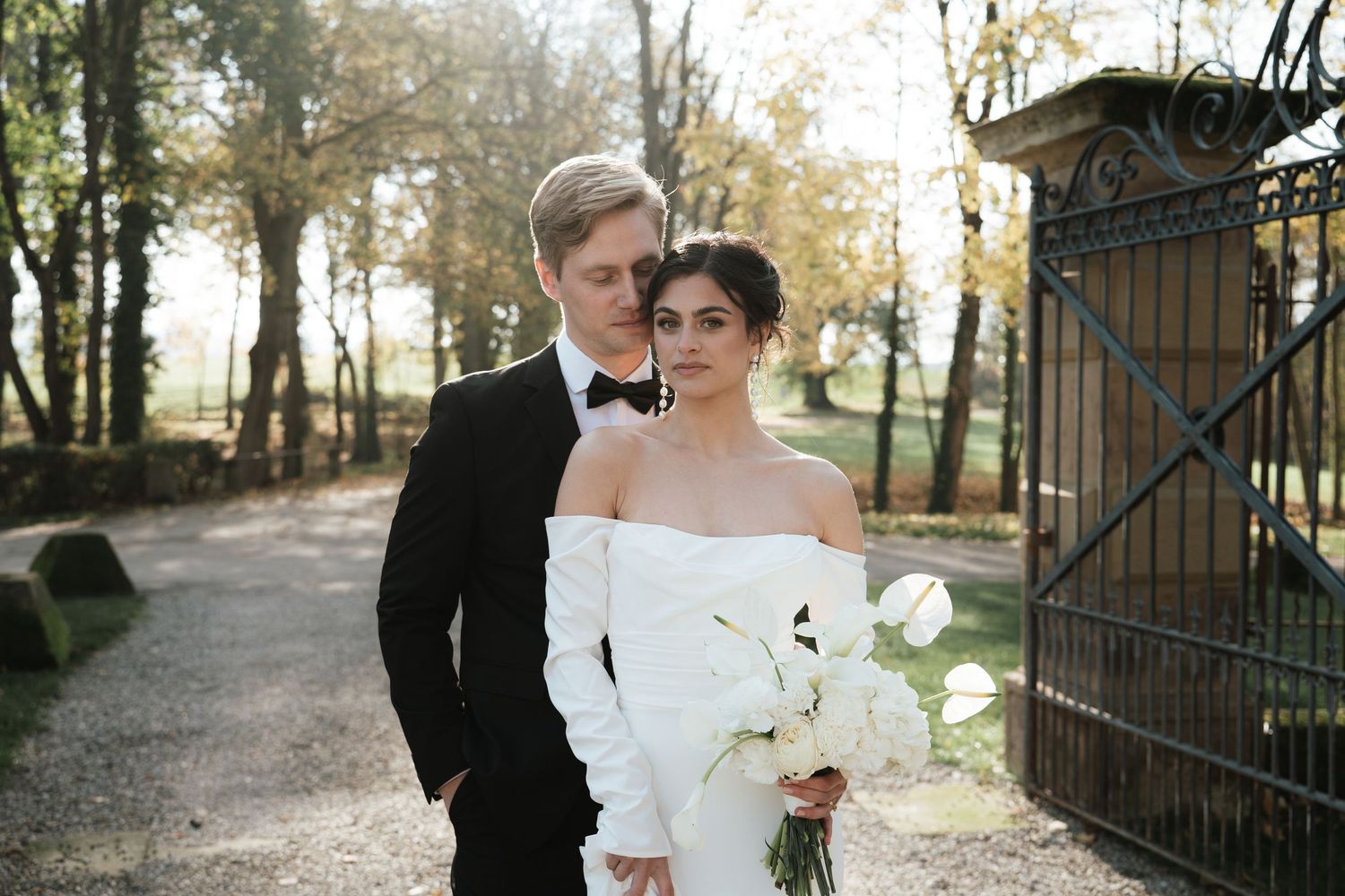 Elegant couple poses in formal wedding attire with white bouquet near rustic garden gate on autumn day.