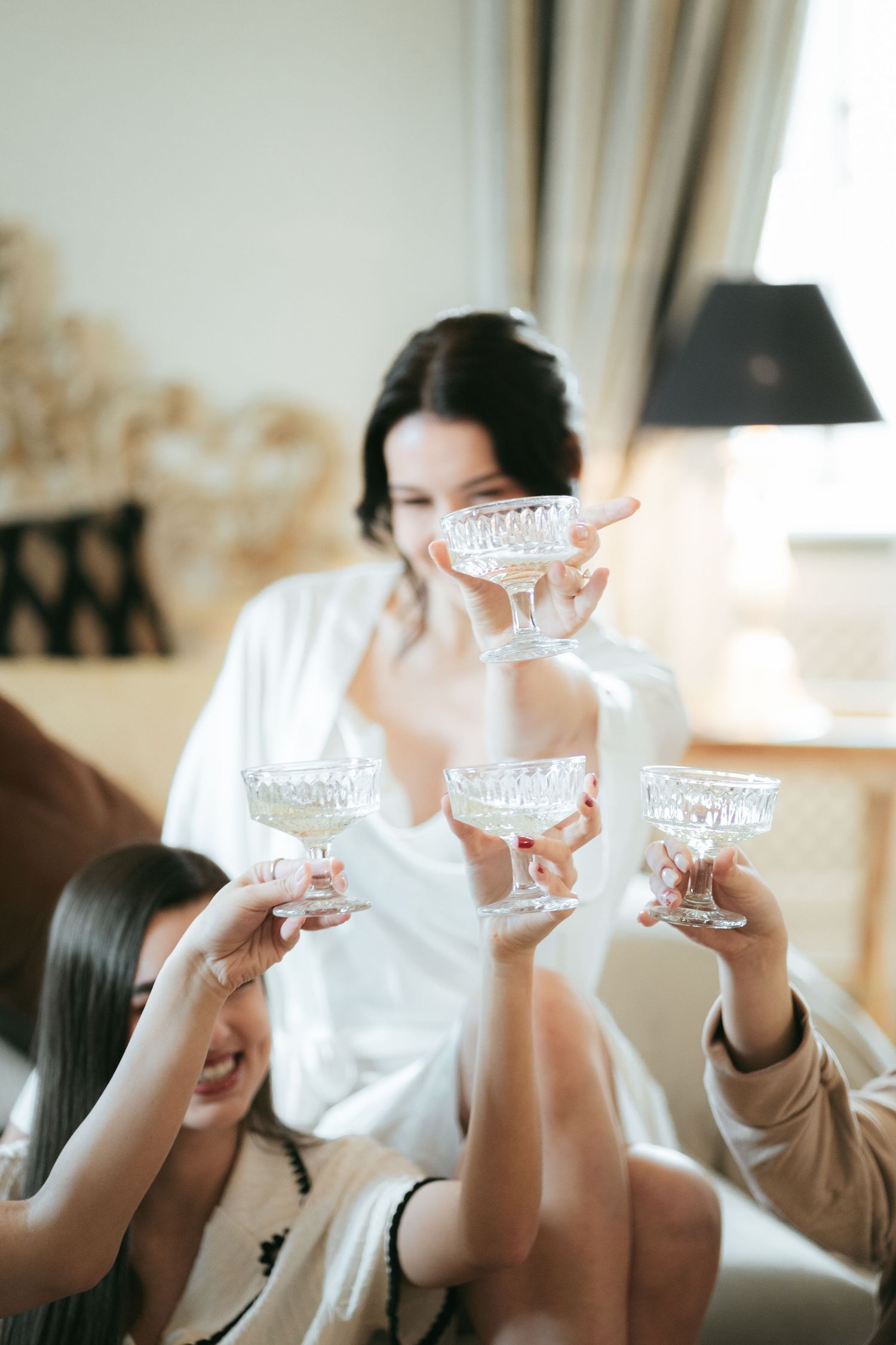 Women in robes toasting with crystal champagne coupe glasses at a bachelorette party celebration indoors.