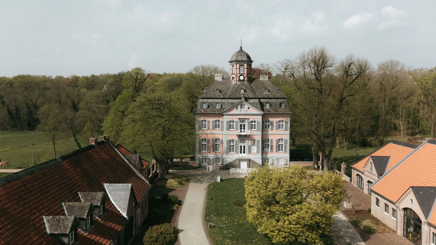 Aerial view of a historic European manor house surrounded by lush trees and traditional farm buildings on a sunny day.