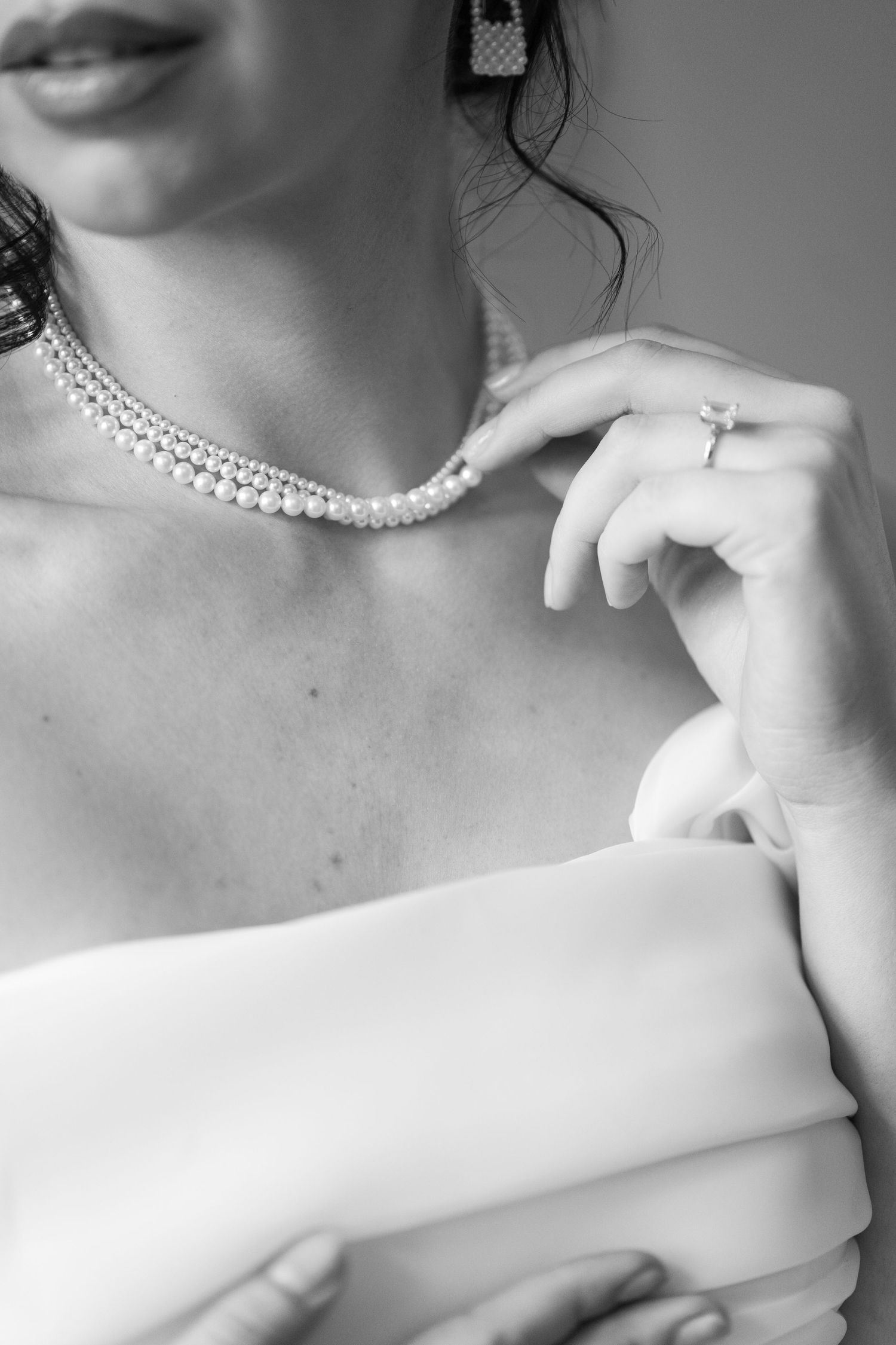 Black and white photo of a bride wearing a pearl necklace and diamond ring in an off-shoulder white gown.