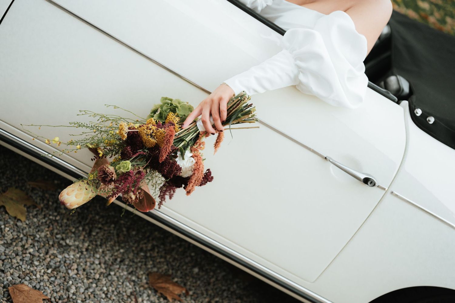 A colorful autumn wedding bouquet rests on the edge of a white vintage car during a fall celebration.