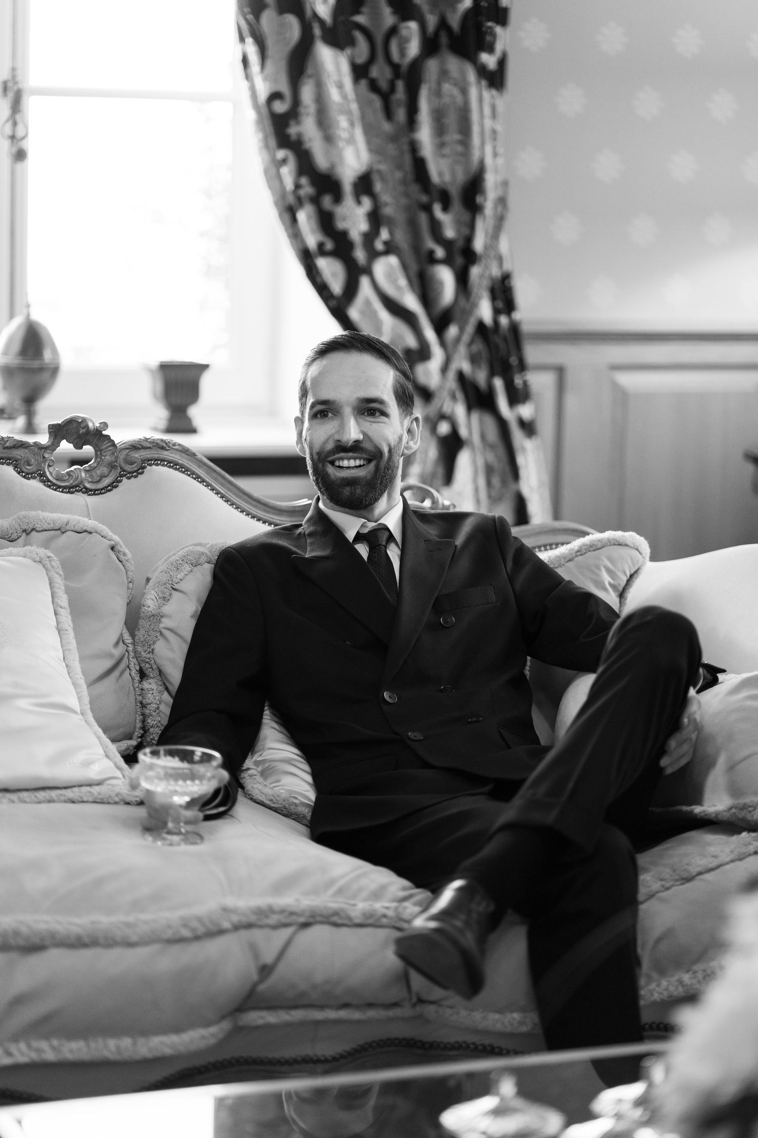 Smiling man in dark suit sitting relaxed on ornate sofa holding drink, elegant room, black and white photo.