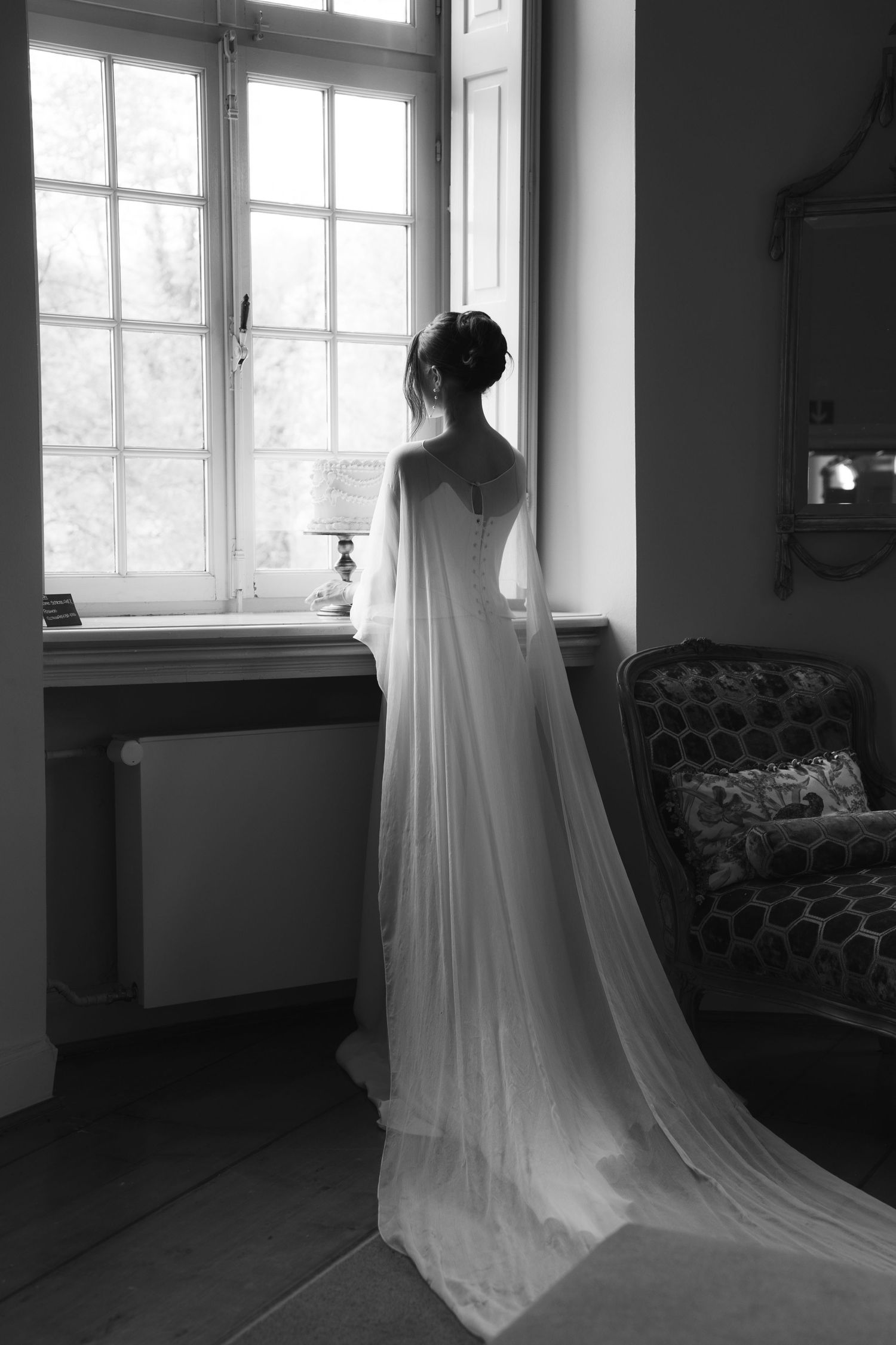 Bride in elegant white wedding gown with flowing cape stands gazing out window in black and white photo.