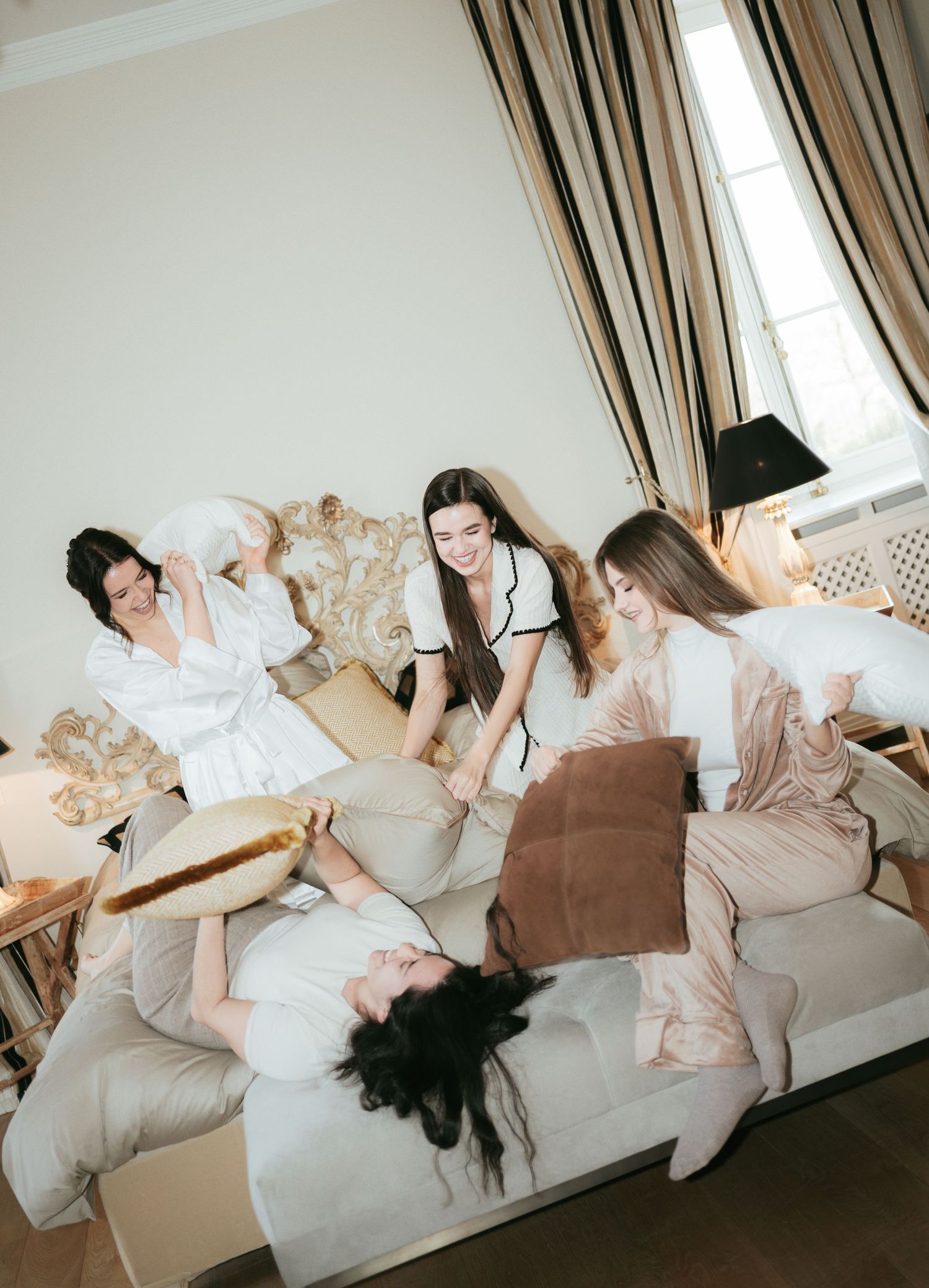 Four women in robes having a playful pillow fight on a couch in an elegant hotel room.