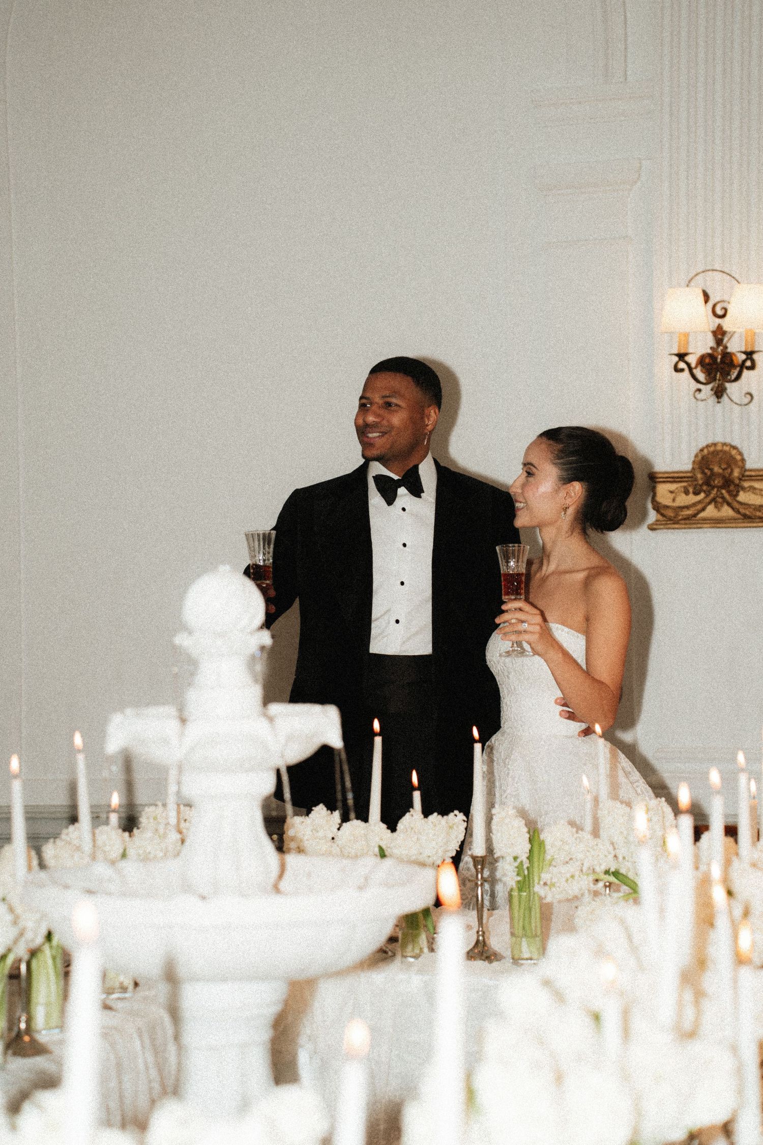 Bride and groom toasting with drinks at elegant candlelit wedding reception with white floral centerpieces.