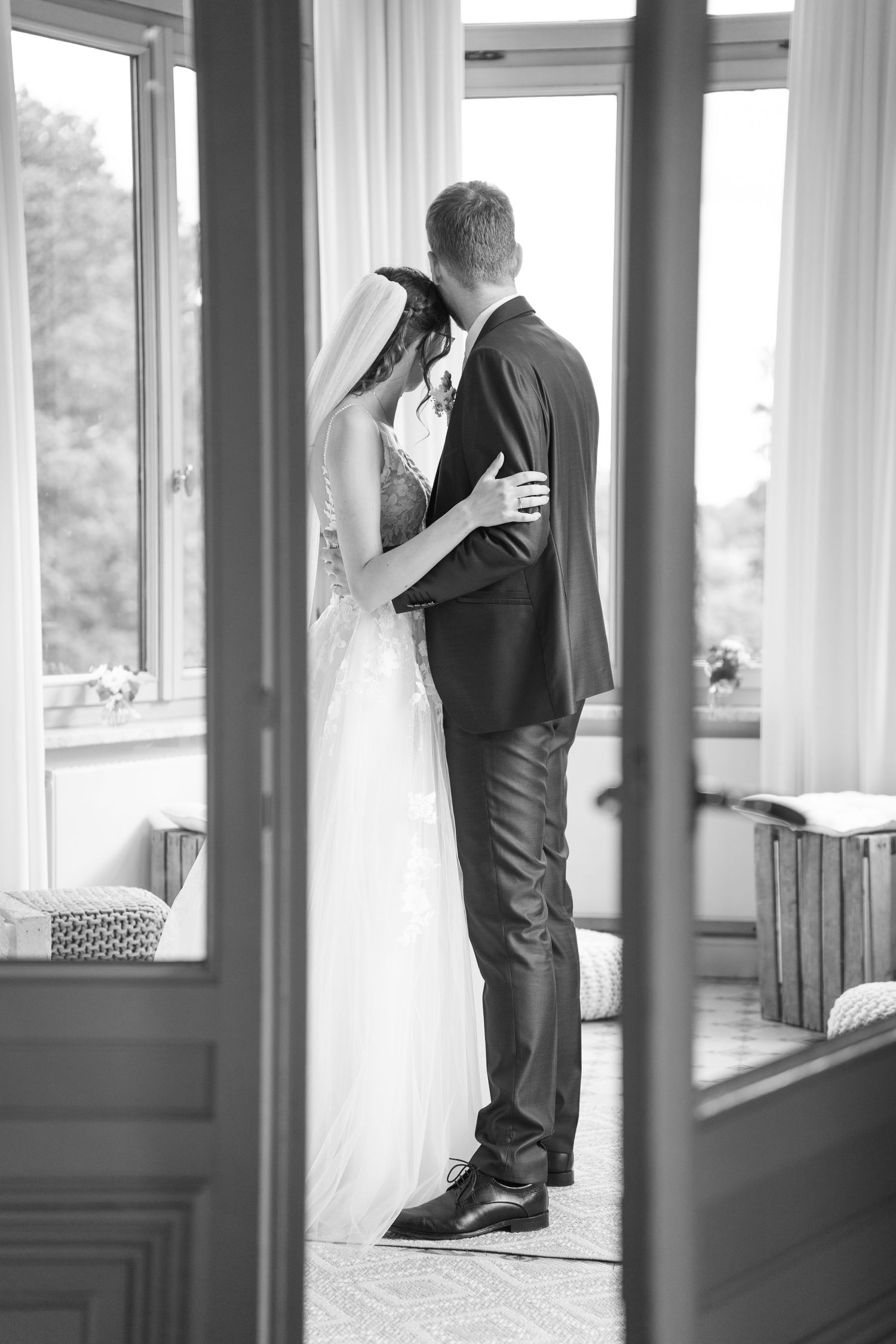 A black and white photo series showing a couple in wedding attire looking out windows in an intimate indoor setting.