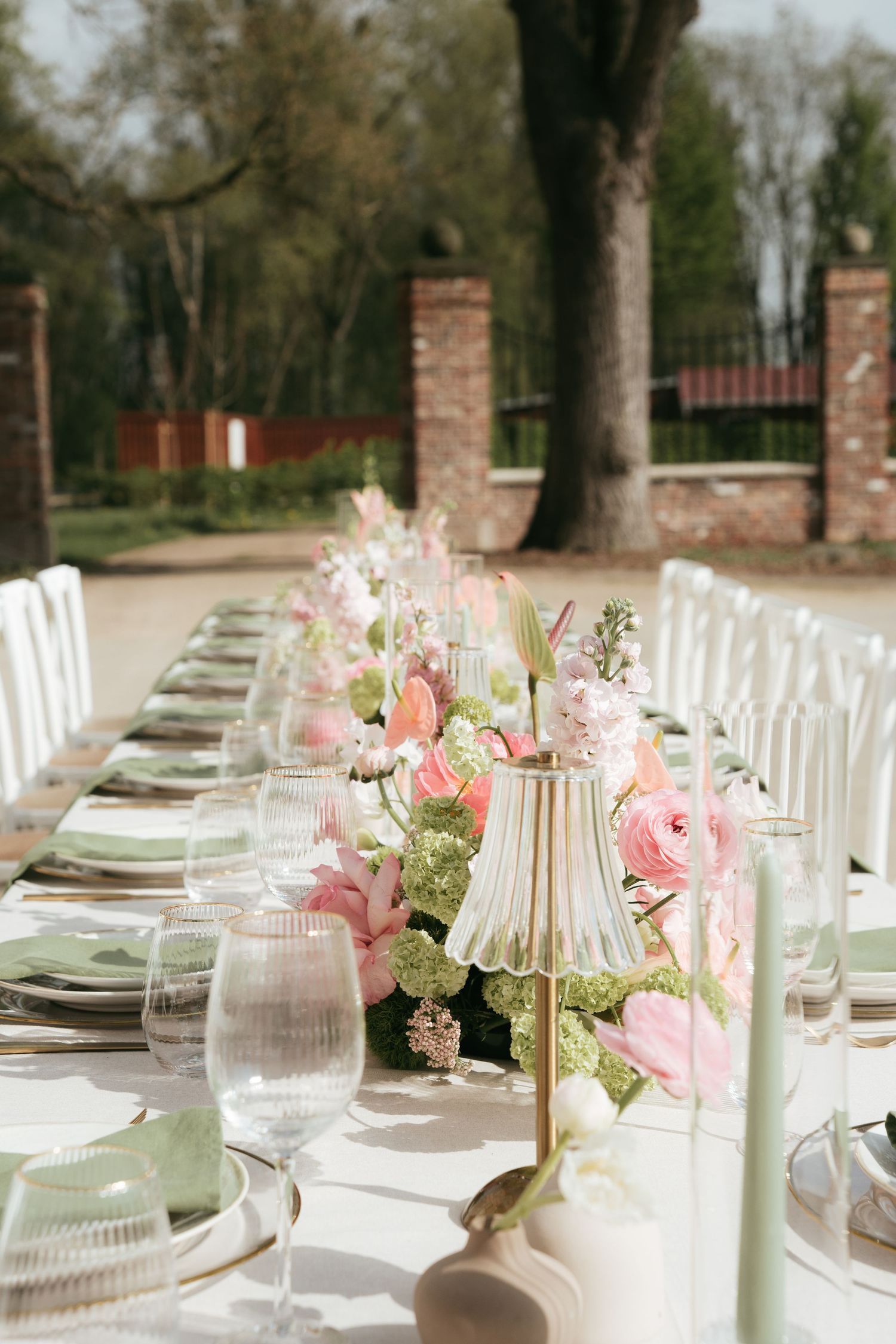 Elegant outdoor wedding reception table with pink floral centerpieces, mint green glassware, and white chairs in a garden setting.