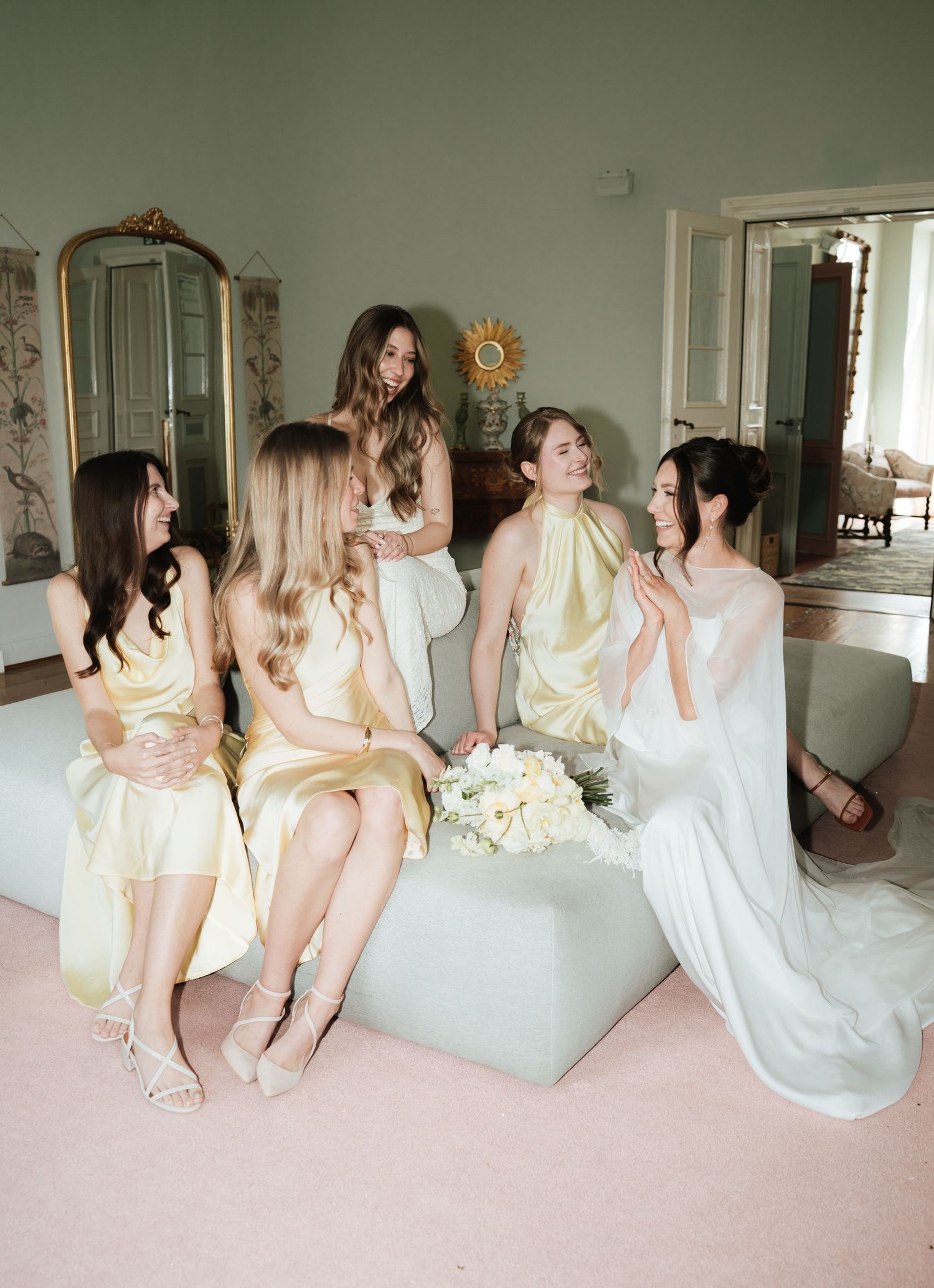 A bride in white holds flowers while chatting with four bridesmaids in yellow dresses in an elegant room.