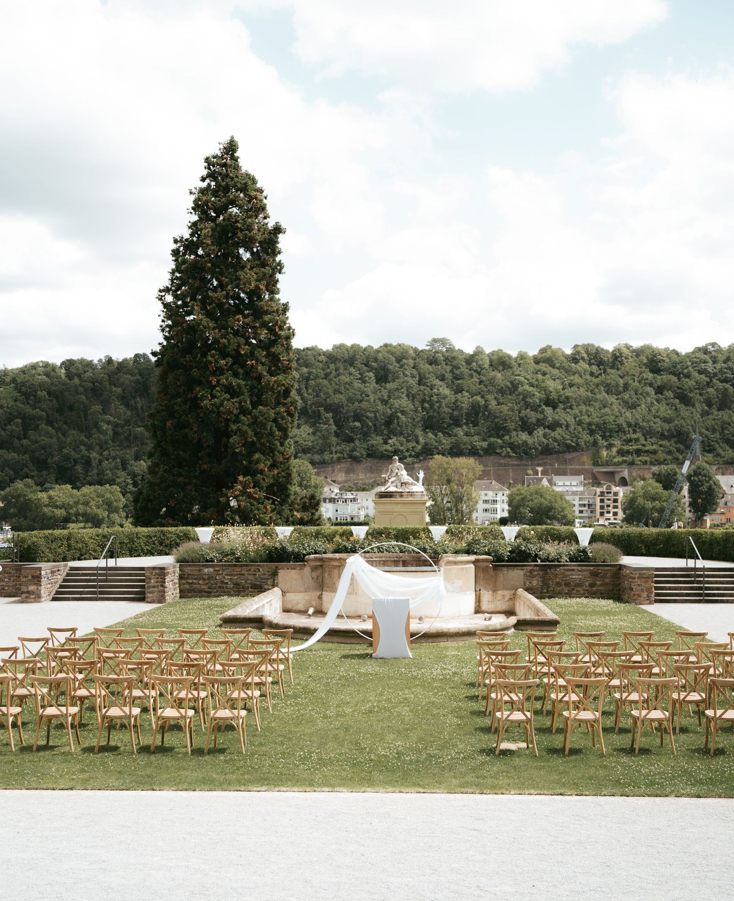 Outdoor wedding ceremony setup with wooden chairs arranged on a grassy lawn facing a rustic wooden arch and floral decor.