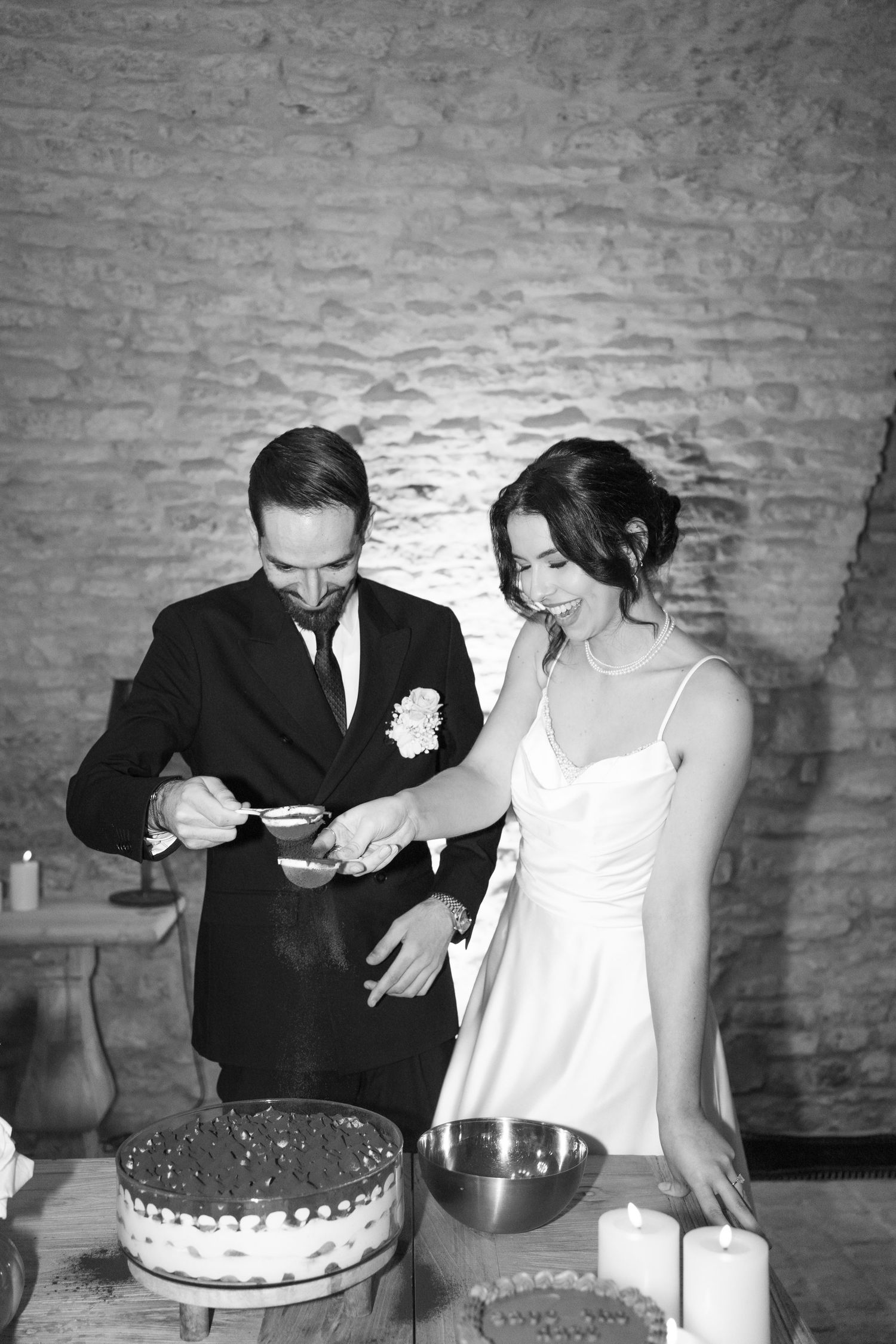 Black and white photo of bride and groom cutting wedding cake in rustic stone venue.