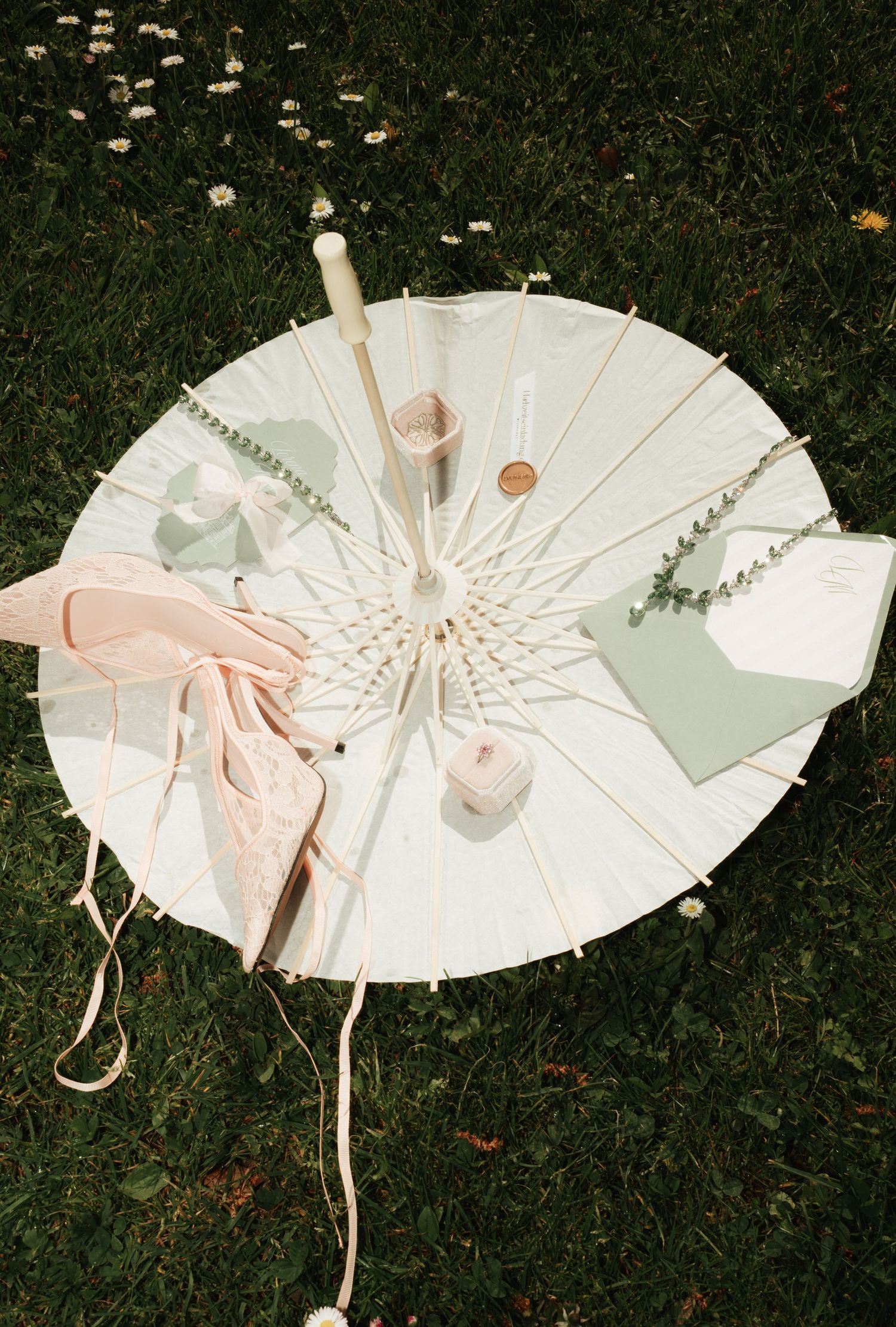 White parasol flat lay on grass with candles, greenery, sage envelopes, and blush pink fabric for a boho wedding.