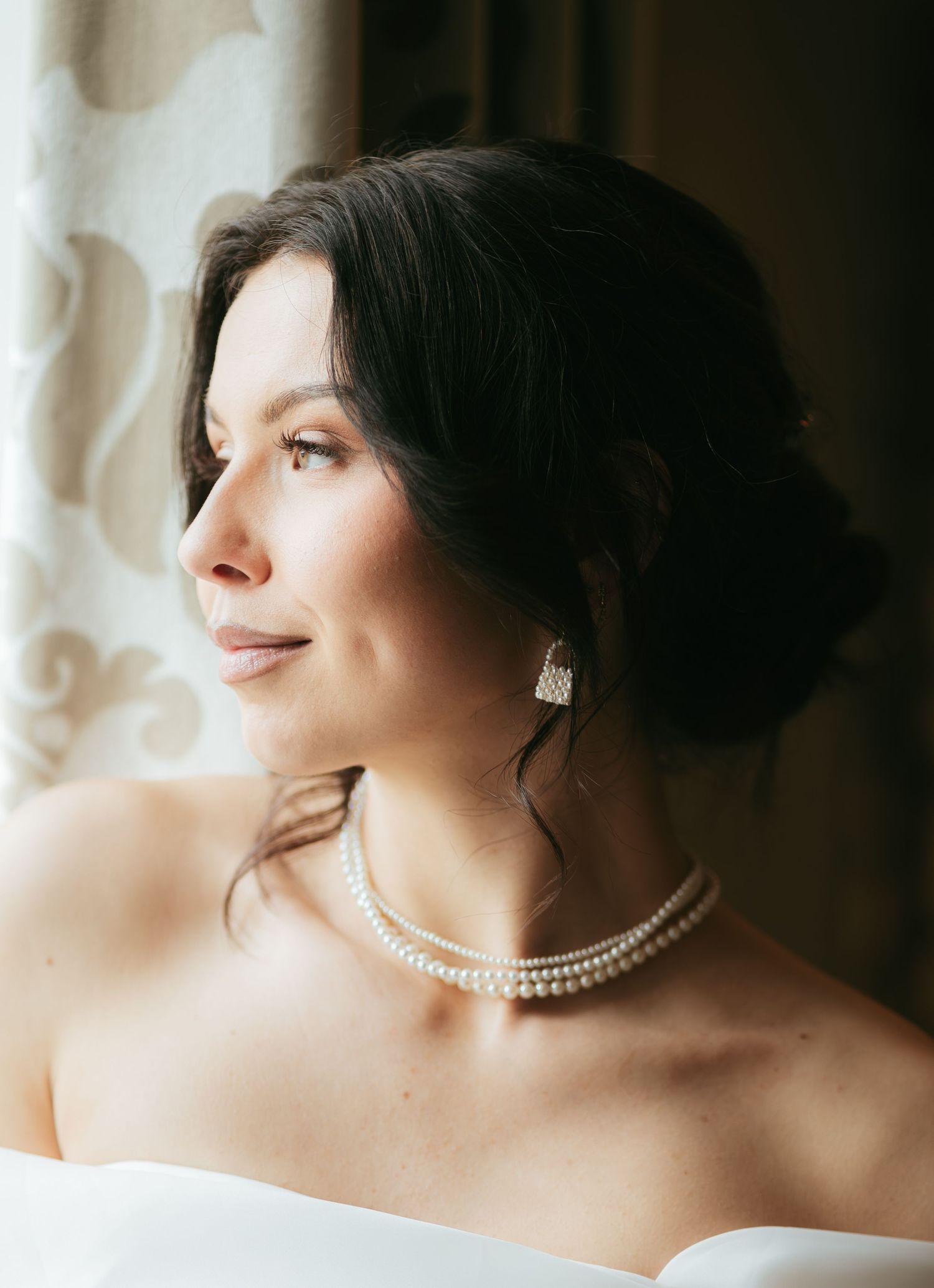 Elegant bride with dark updo hair, pearl necklace and earrings, wearing strapless white gown, gazing softly toward window light.