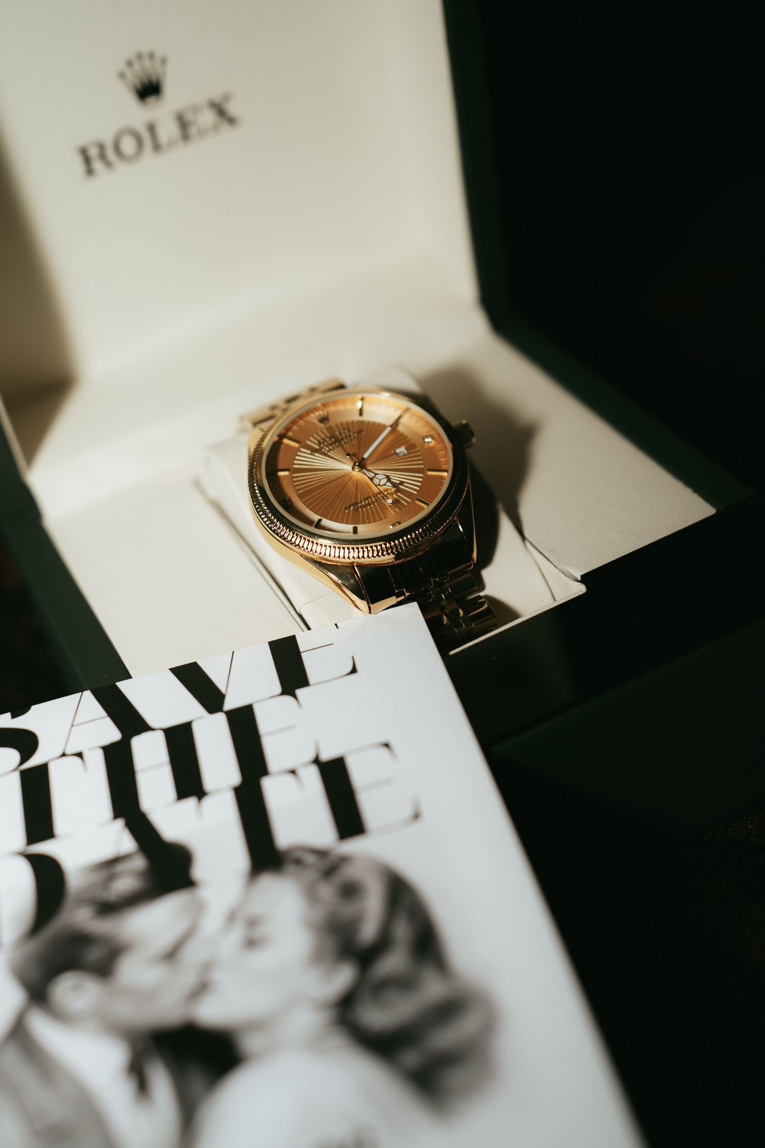 Gold Rolex watch in luxury box displayed on a Save the Date wedding card with romantic couple photo.