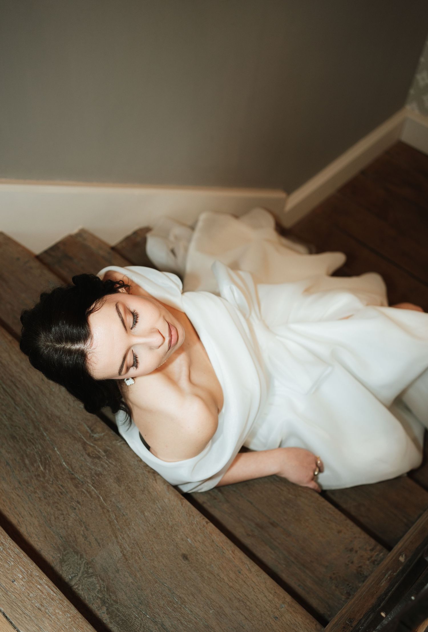 Bride in white gown lying gracefully on rustic wooden staircase, smiling softly with eyes closed.
