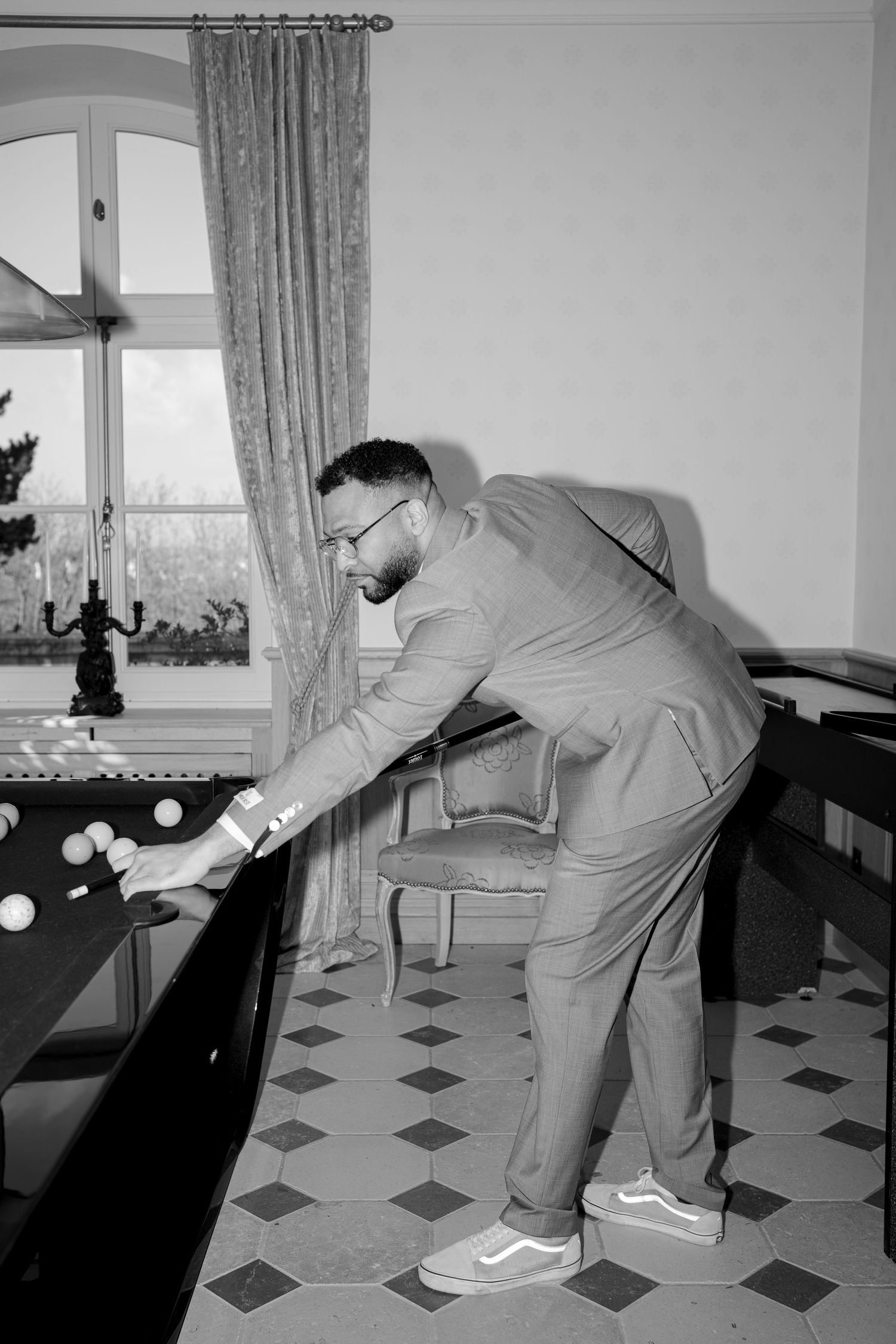 Man in suit and sneakers playing billiards in an elegant room with checkered floors and draped windows.
