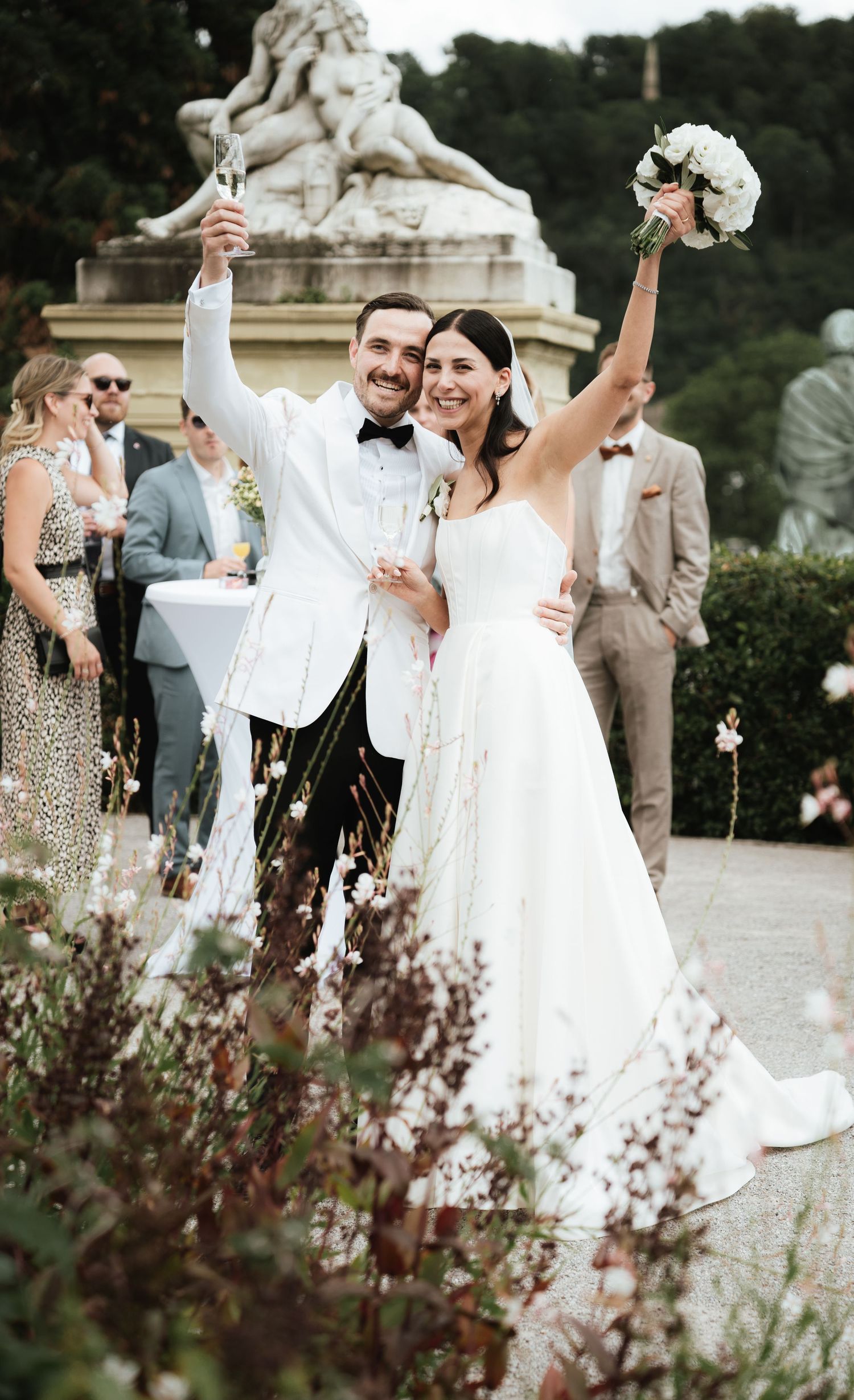 Wedding party celebrates joyfully outdoors in a garden setting with raised arms and bouquets.