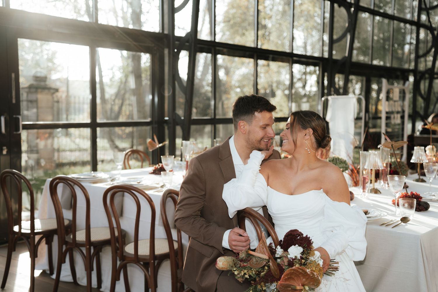 Newlyweds share an intimate moment at their reception in a glass-enclosed venue with rustic wooden chairs and floral decor.