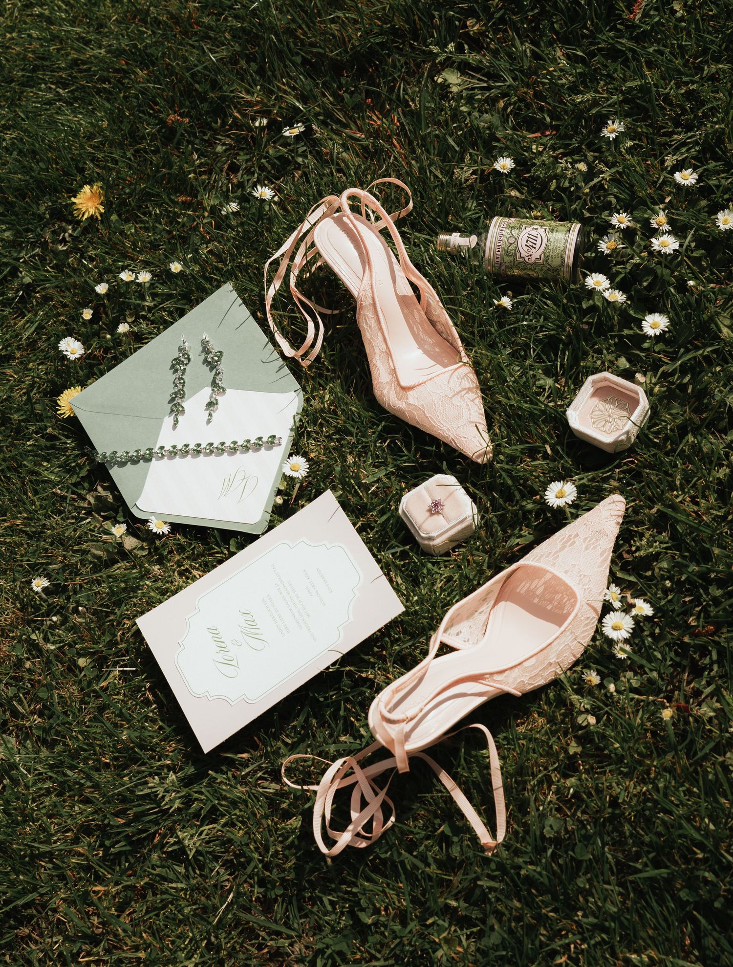 Blush pink lace heels, sage green wedding invitations, earrings, and ring box arranged on grass with daisies.
