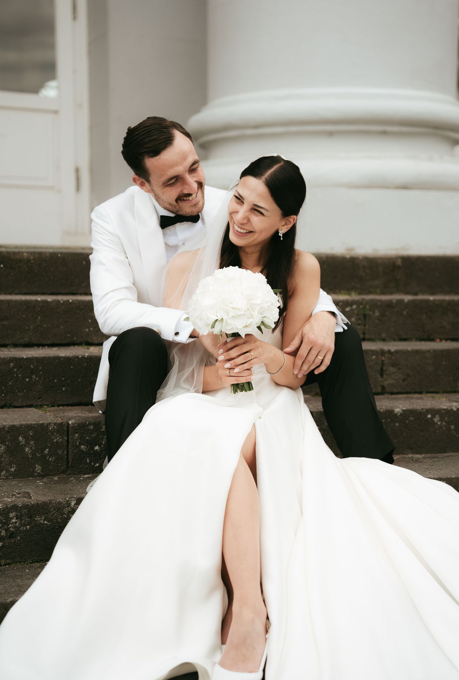 A couple sits together on stone steps, with the bride in a white gown holding a bouquet and groom in white tuxedo jacket.