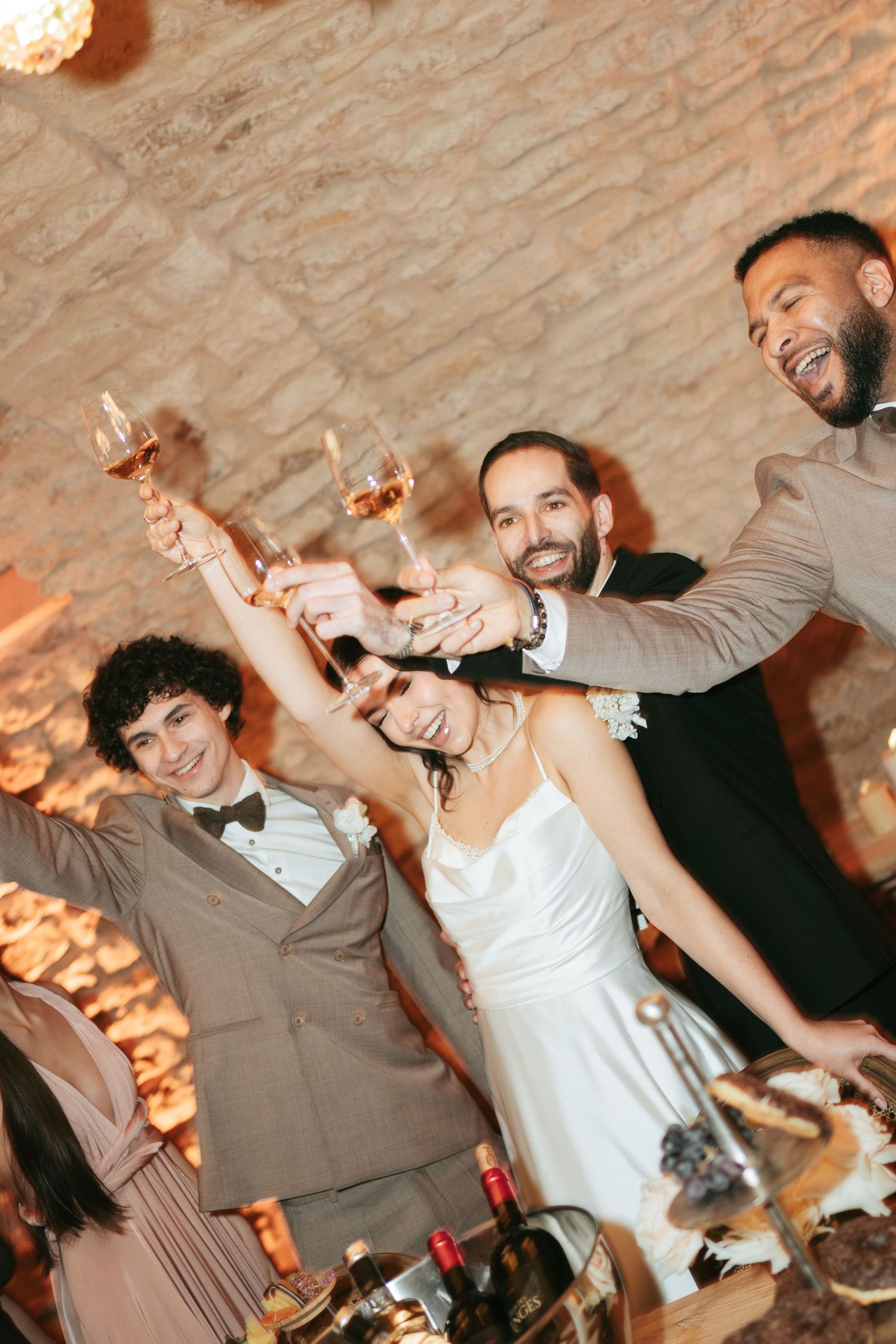 A joyful wedding toast with bride in white dress and guests raising champagne glasses at a rustic stone venue.