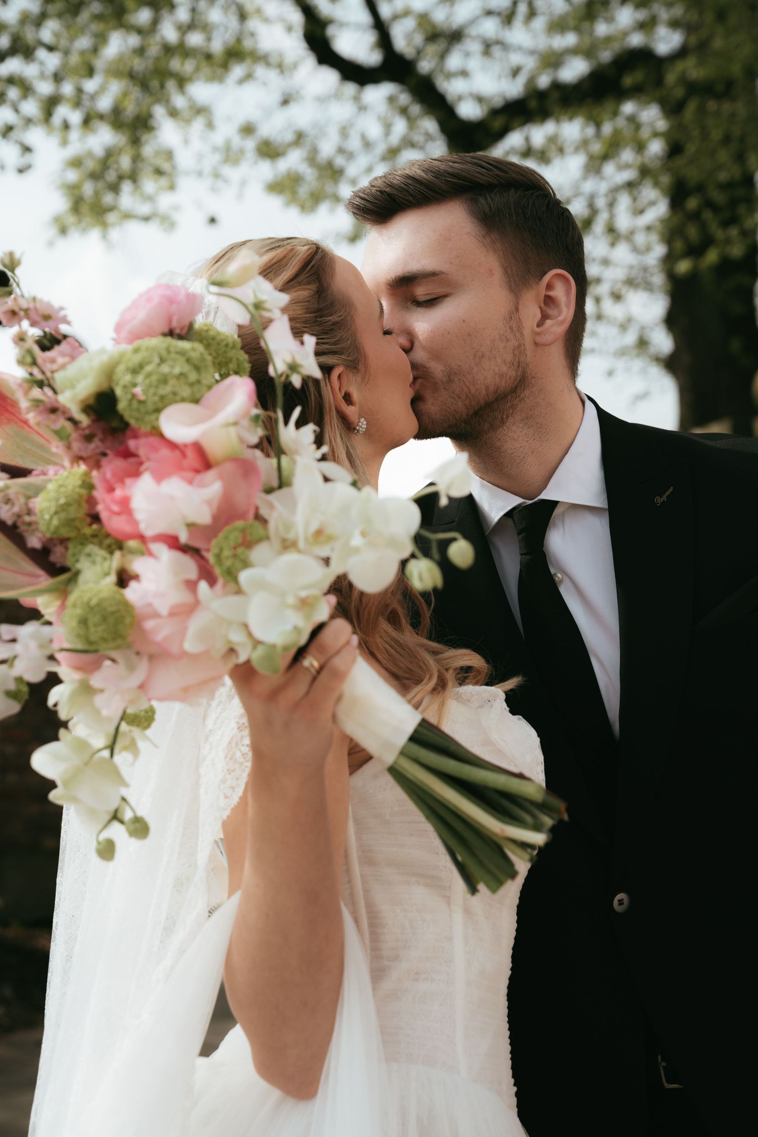 Bride and groom sharing a kiss on their wedding day, with a beautiful pink and white floral bouquet in the foreground.