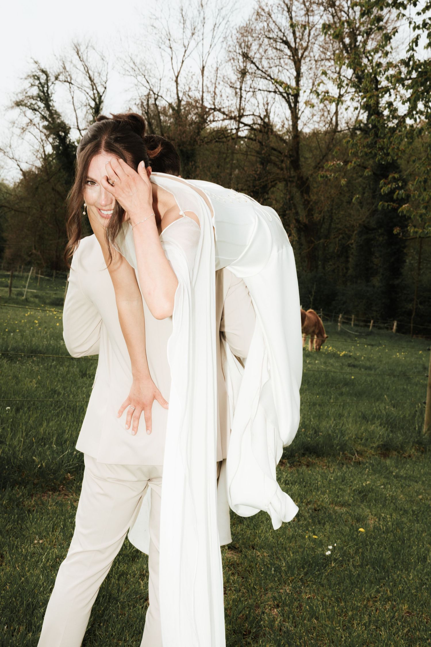 Smiling bride in white suit carries wedding dress over shoulder in lush green field with trees and horse in background.