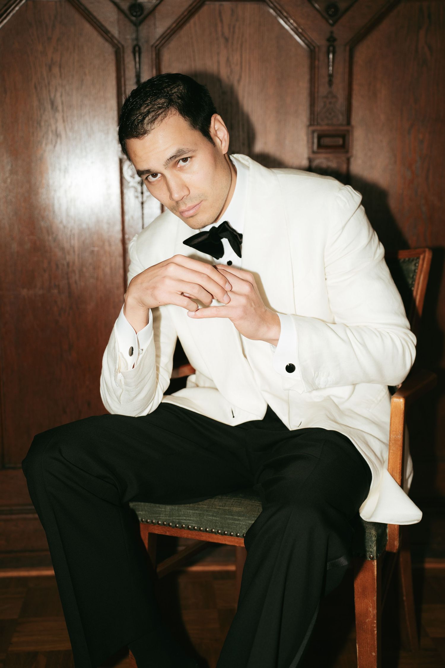 A man in a white tuxedo jacket and black bow tie sits confidently on a wooden chair with hands clasped.