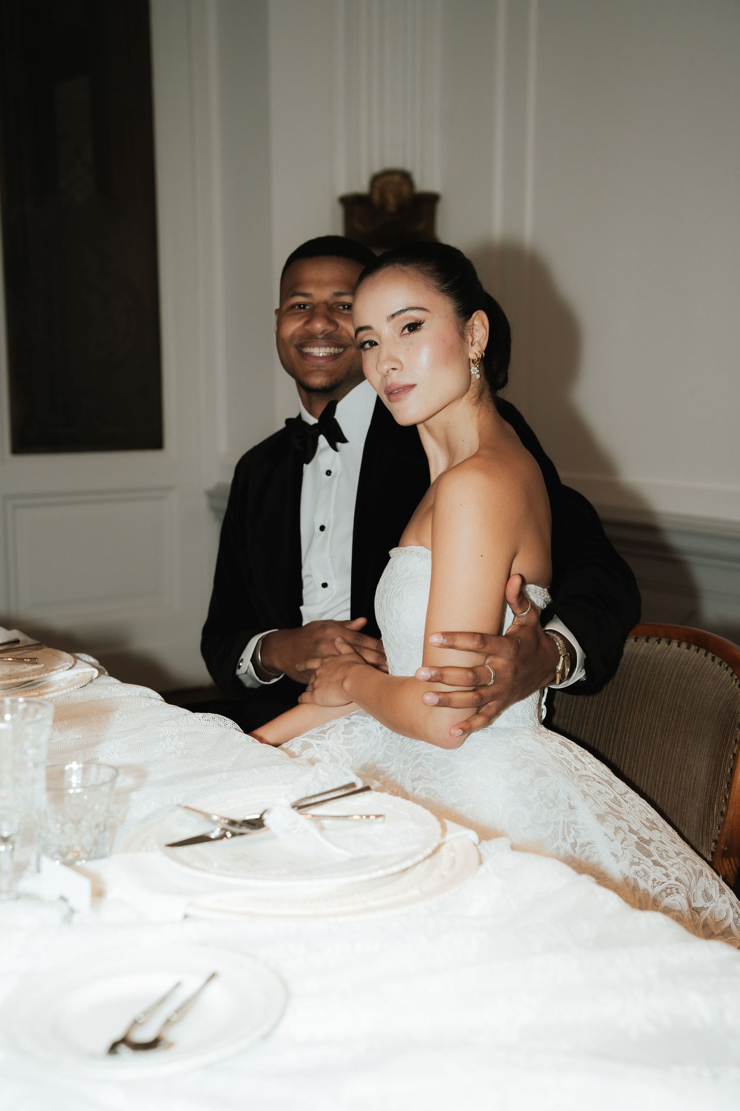 Bride and groom seated at elegant wedding reception table, groom in tuxedo embracing bride in strapless lace gown.