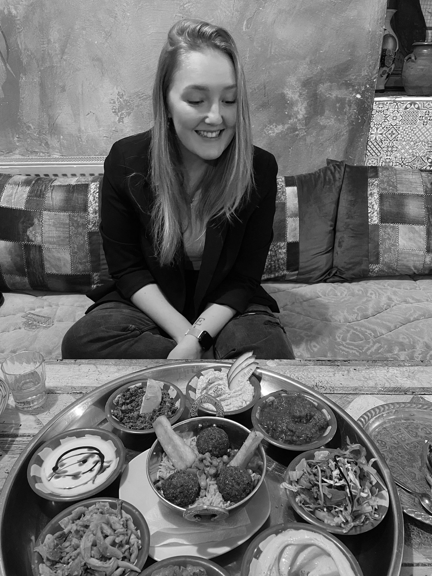 Woman smiling at a large tray of Middle Eastern dishes in a cozy restaurant setting, black and white photo.