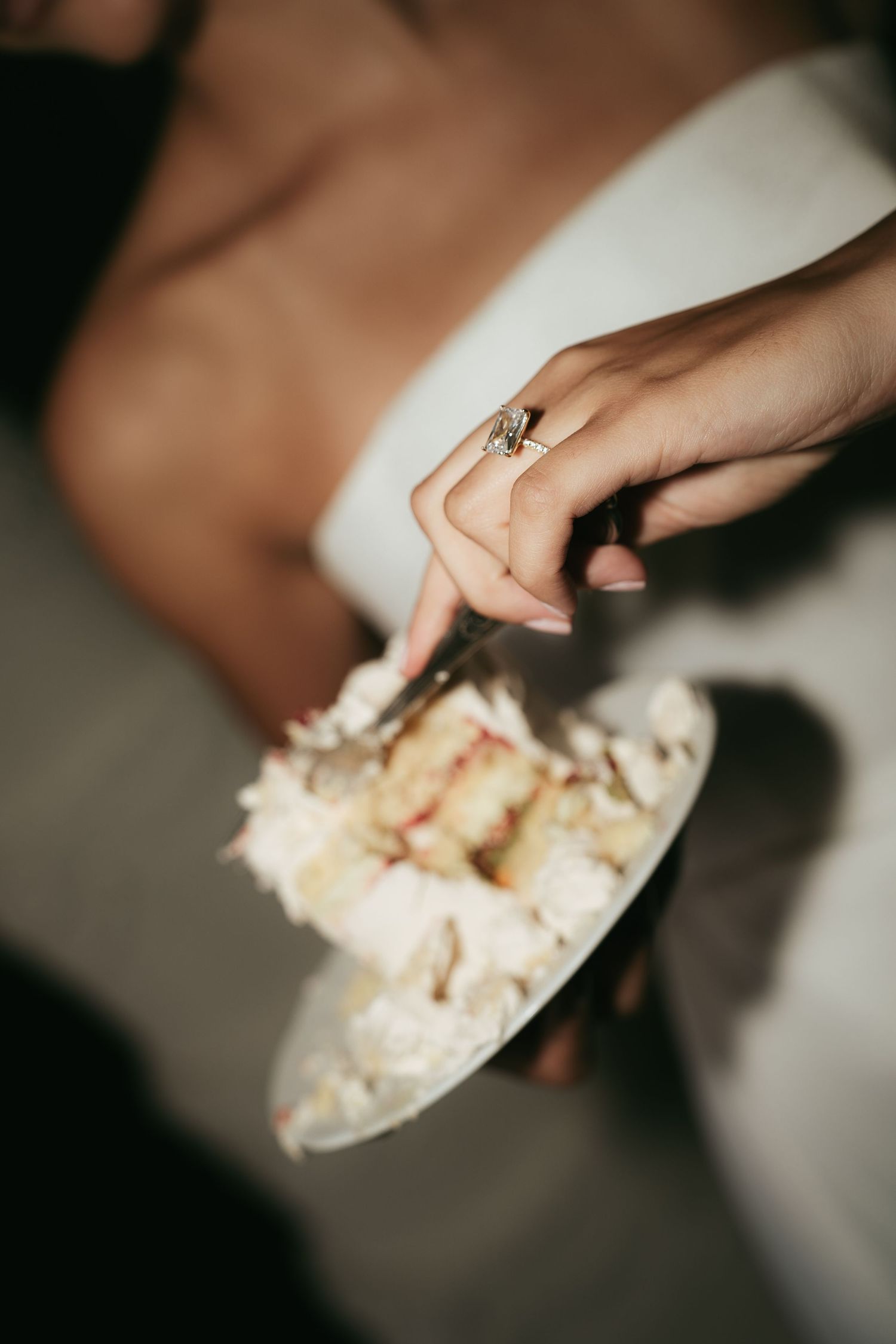 Bride wearing gold diamond ring holding a plate of white wedding cake with a fork.