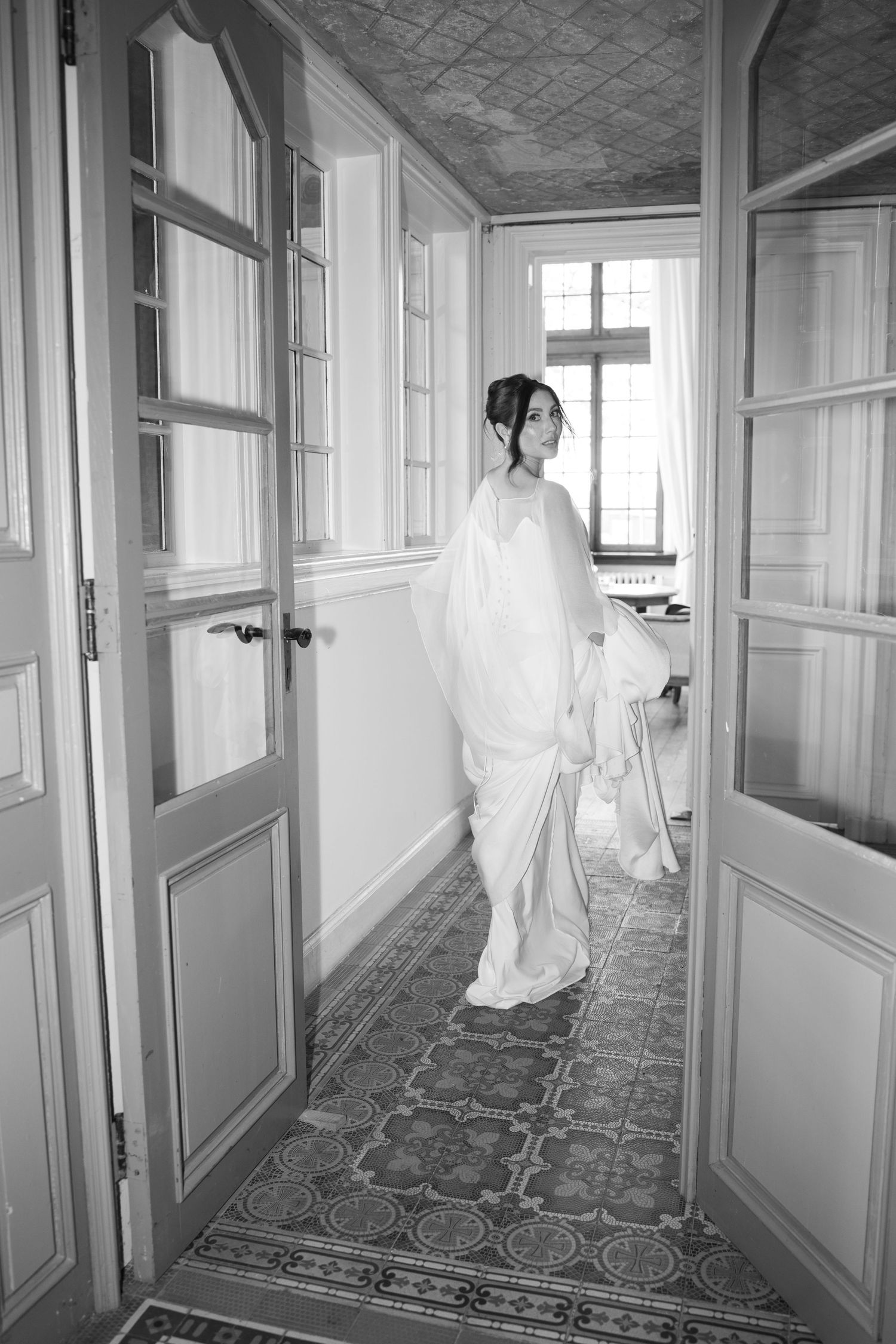 Black and white photo of a bride in a white gown standing in an elegant hallway with ornate tiled floors.