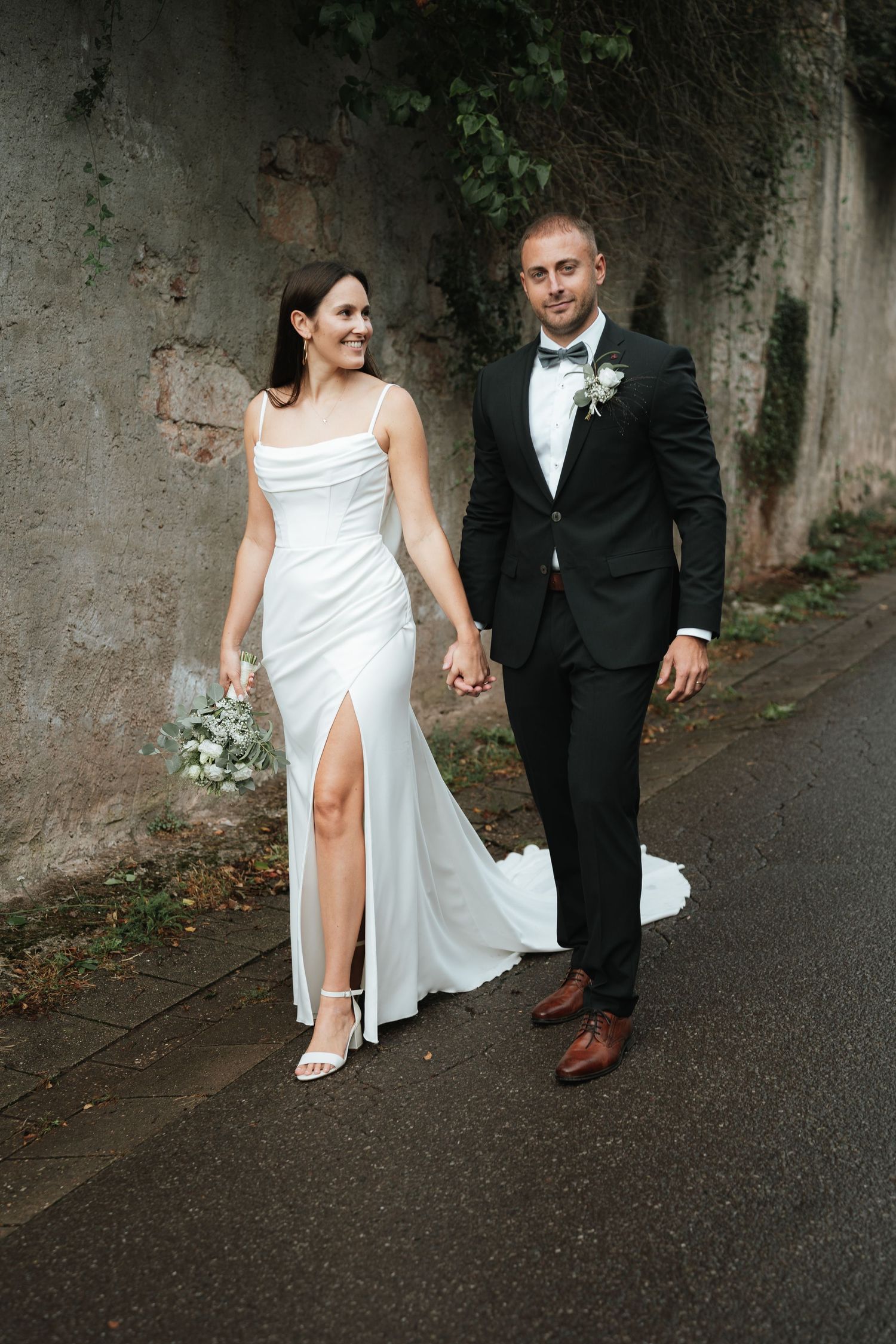A couple in wedding attire walks together on a dark paved path with a stone wall in the background.