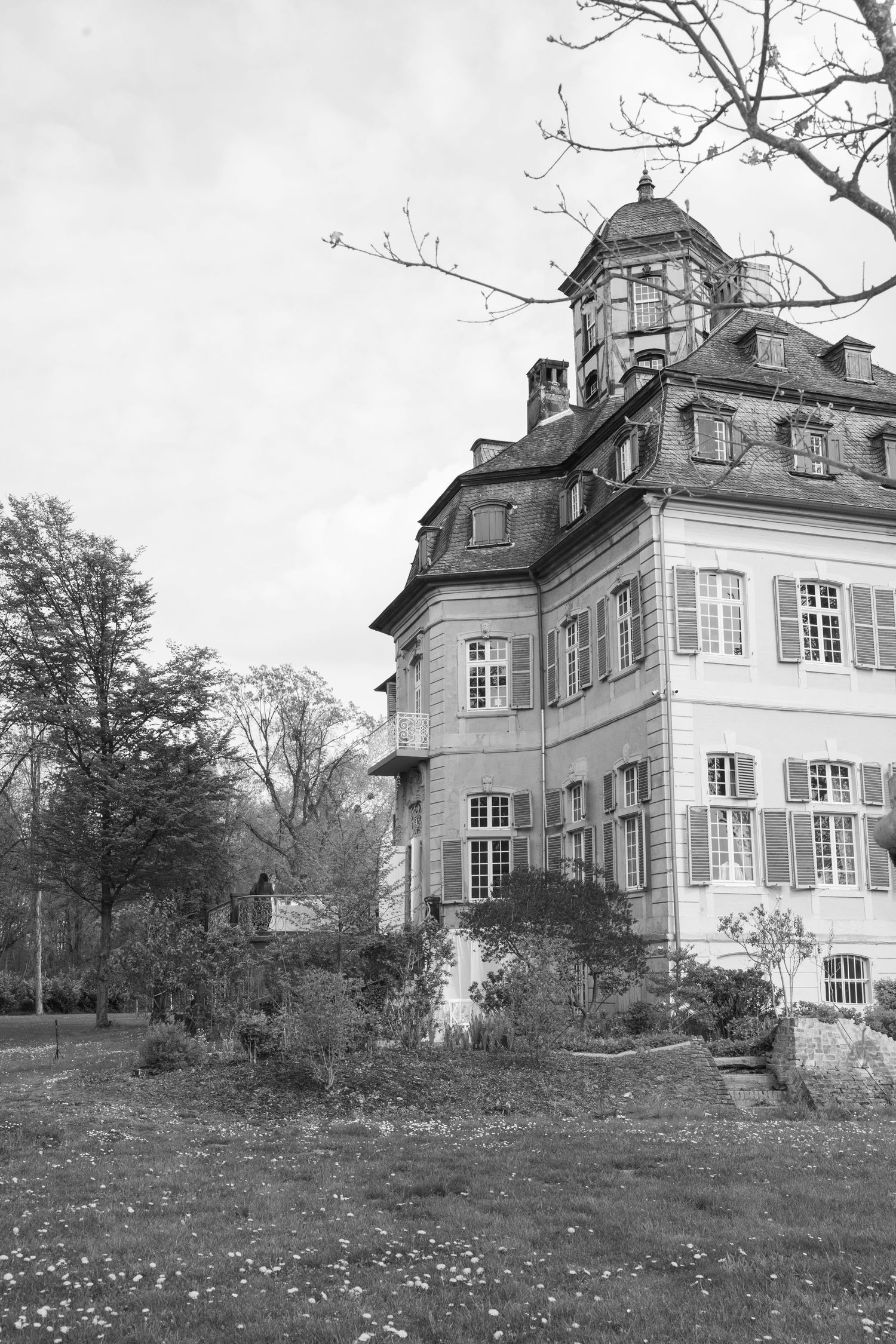 Black and white photo of a historic European mansion with shuttered windows, ornate tower, and lush garden grounds.