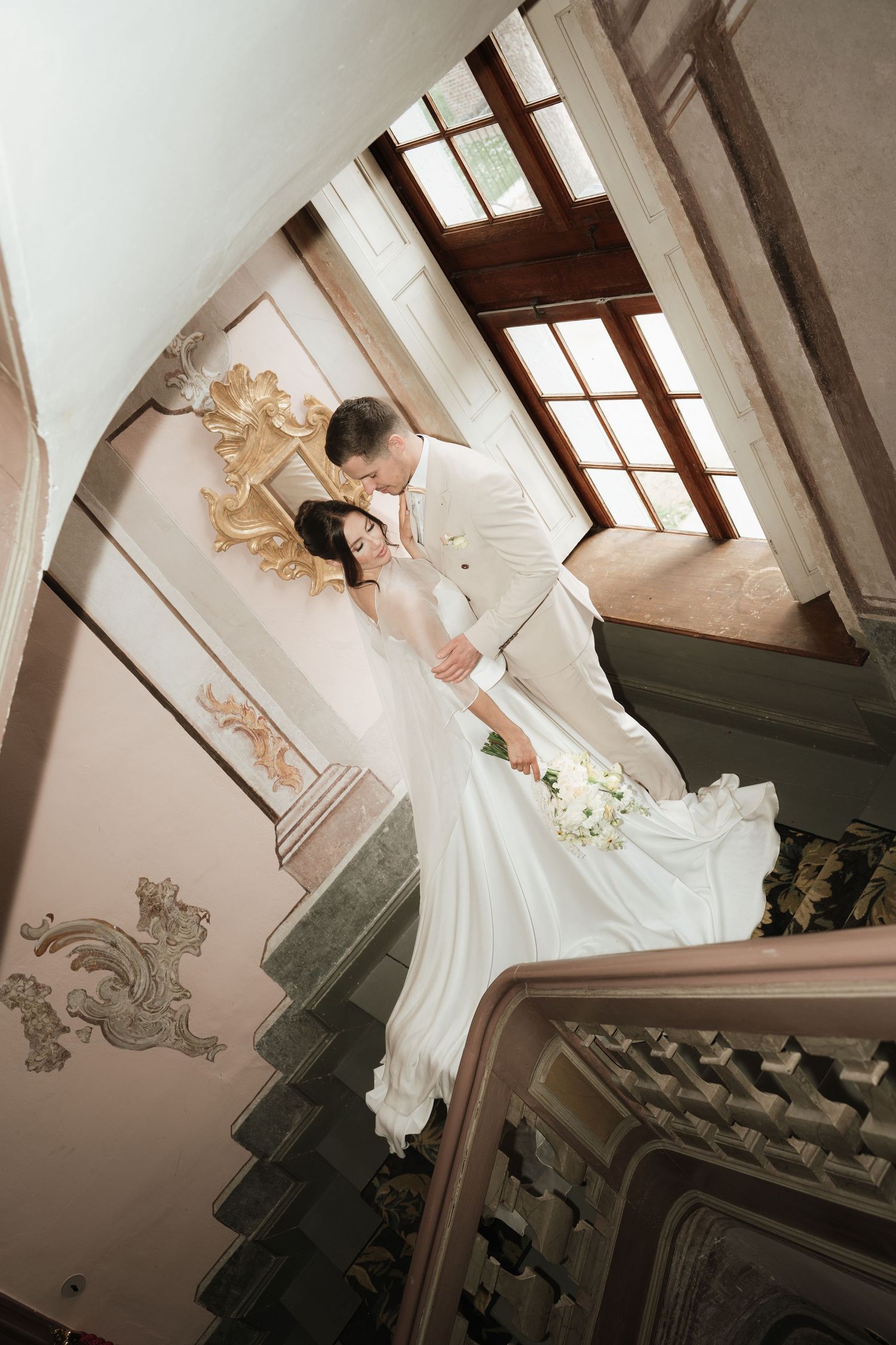 Bride in white gown and groom in white suit share romantic moment on ornate staircase in elegant venue.