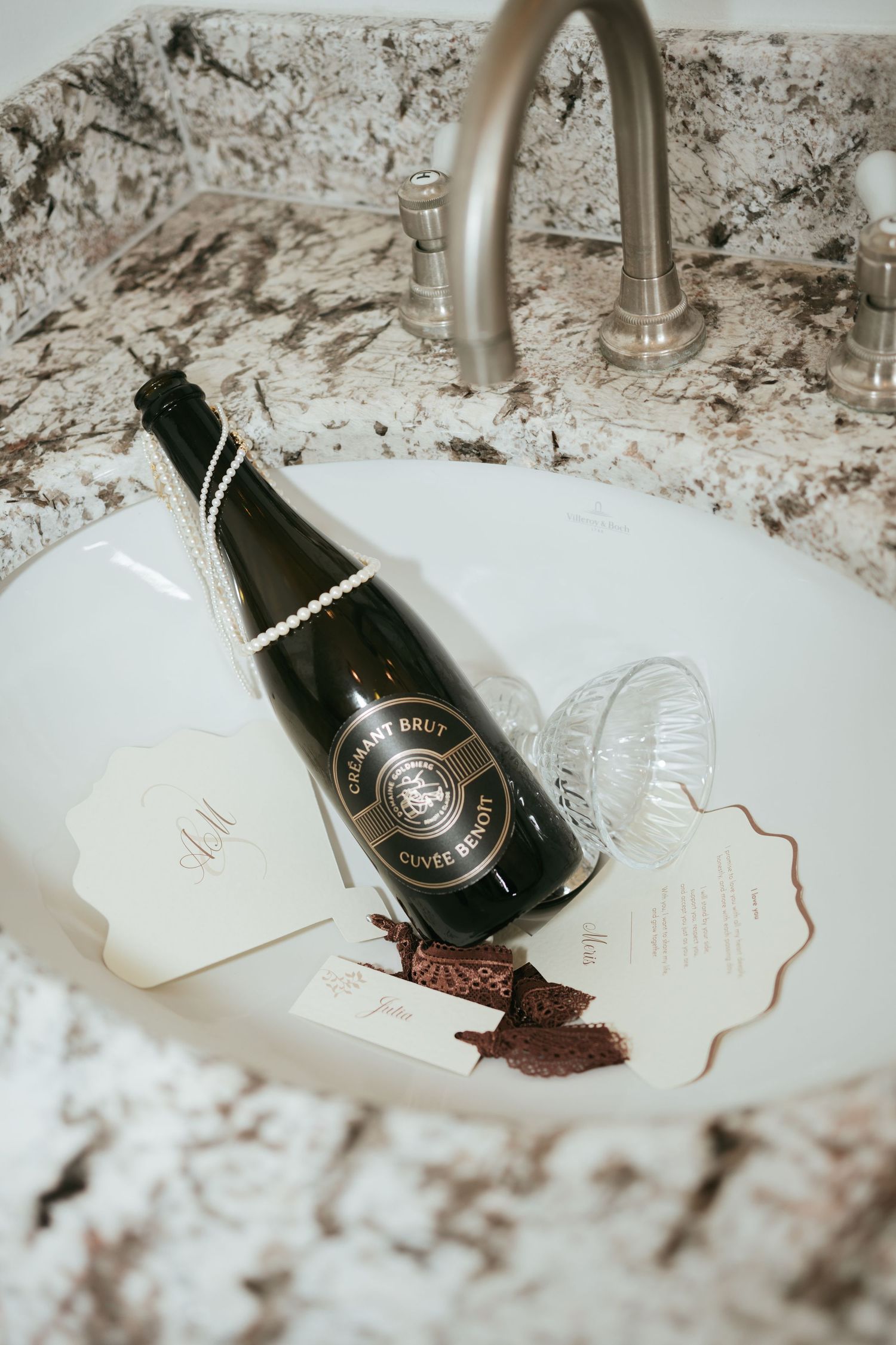 Champagne bottle with pearl necklace and wedding stationery resting in a marble sink basin.