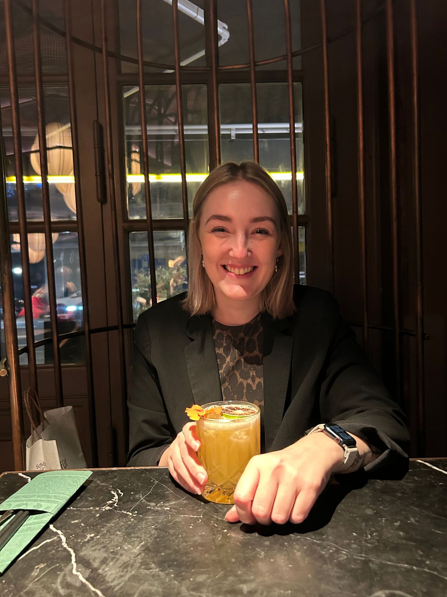 Smiling woman holding a golden cocktail in a mug at a dark bar table, wearing a black blazer.