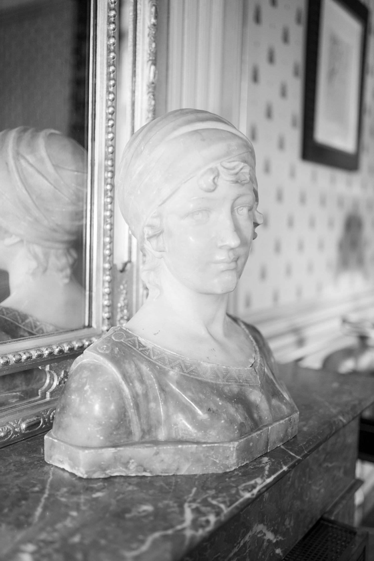 Black and white photo of a marble female bust sculpture on a mantle reflected in an ornate gold mirror.