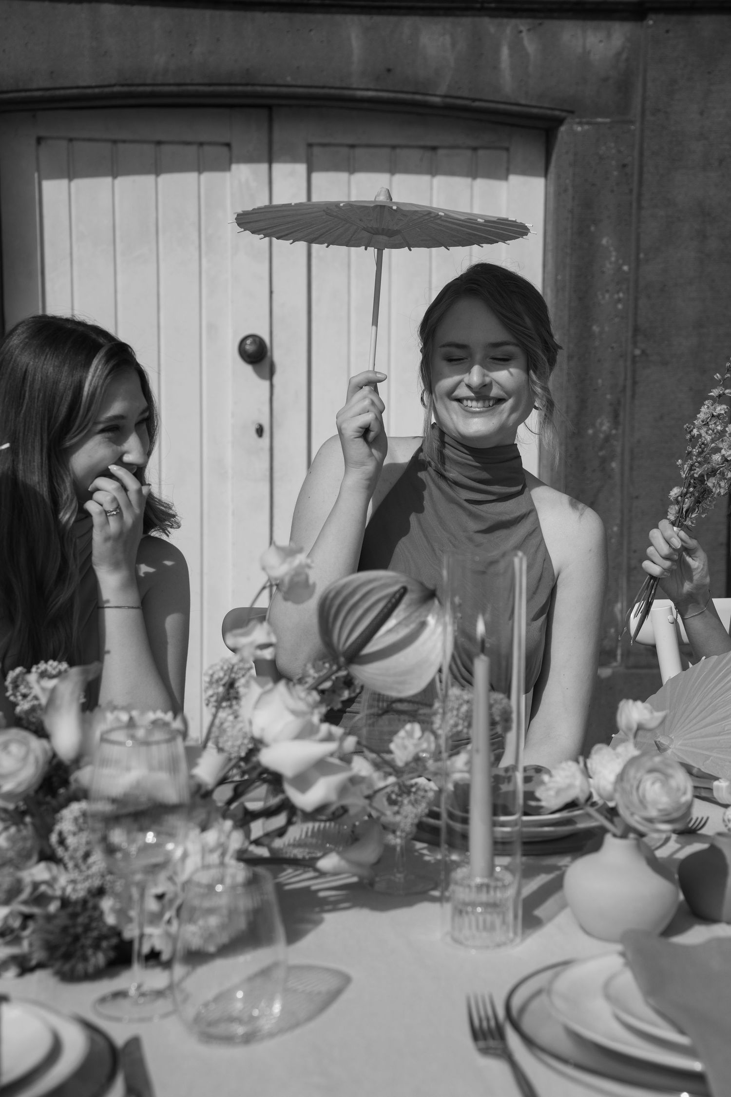 Black and white photo of two women laughing at an elegantly decorated wedding table with flowers and candles.