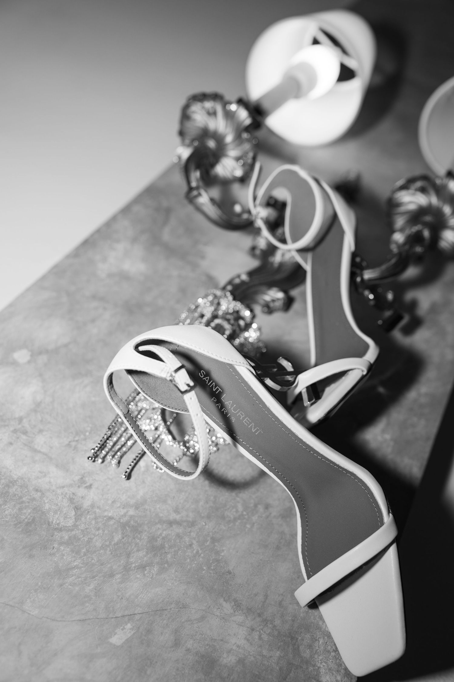Black and white photo of elegant strappy heeled sandals on a marble surface with jewelry and a lamp in the background.