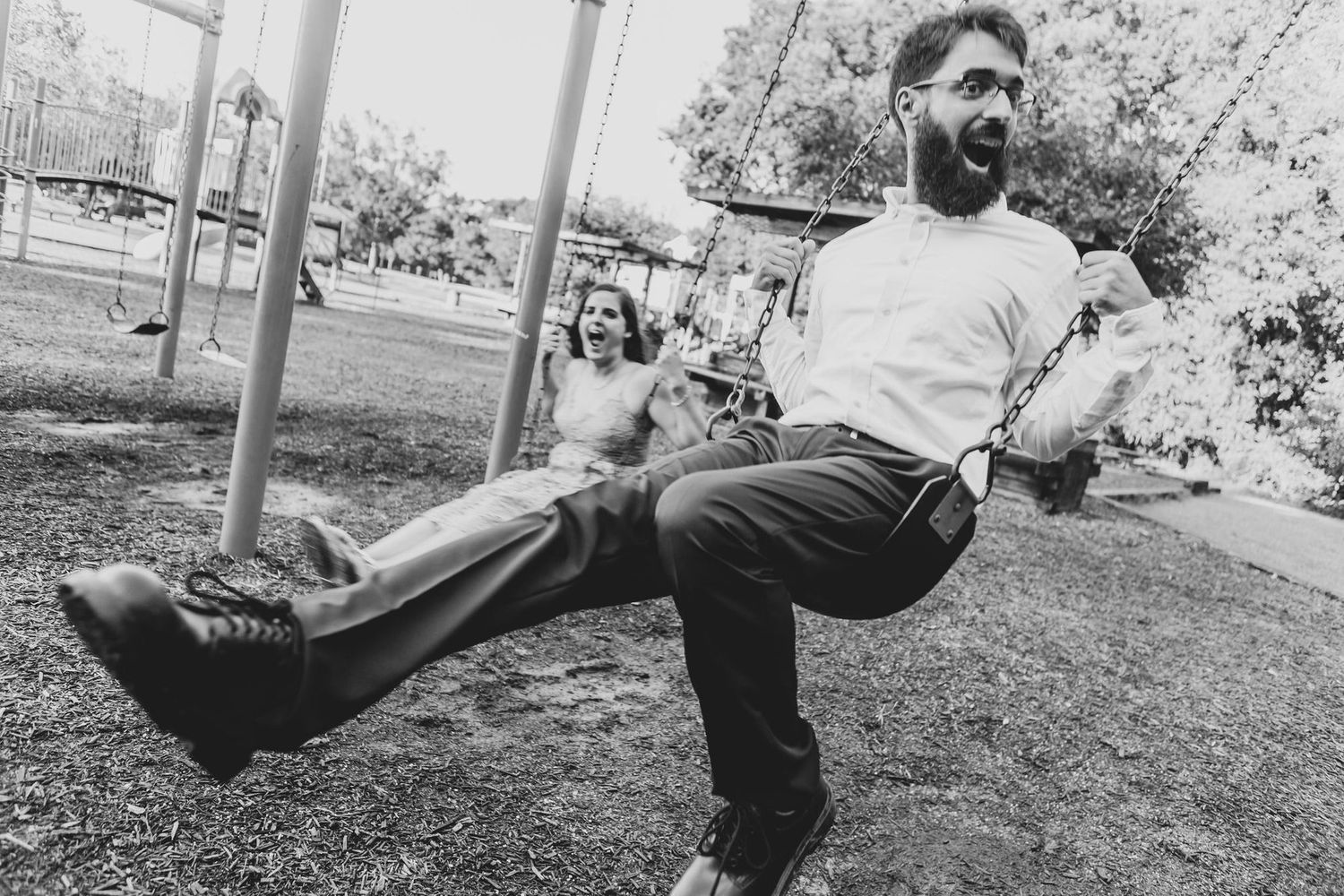 Black and white photo of a bearded man on a swing kicking his legs up at a playground, with a woman swinging behind him.