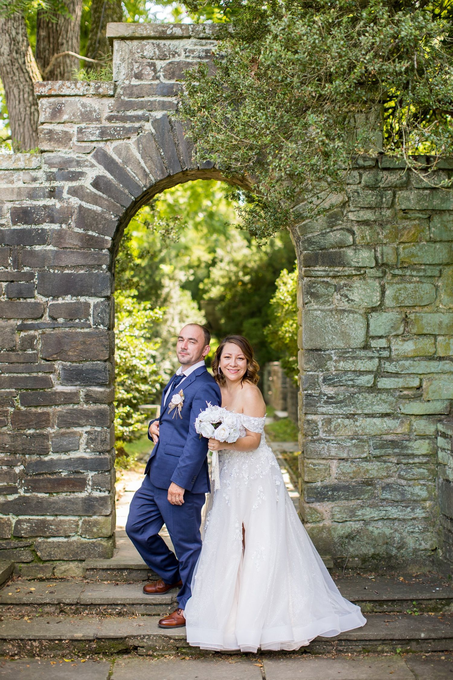 Bride and groom posing together under a stone arch gateway surrounded by lush green trees on their wedding day.