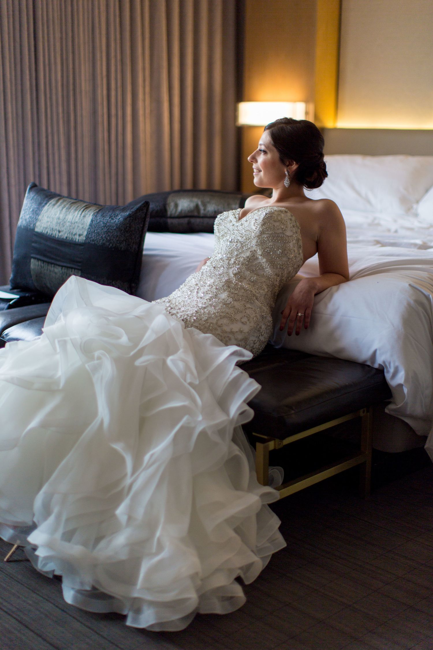 Bride in a white ruffled wedding gown sitting elegantly against a hotel bed, gazing thoughtfully to the side.