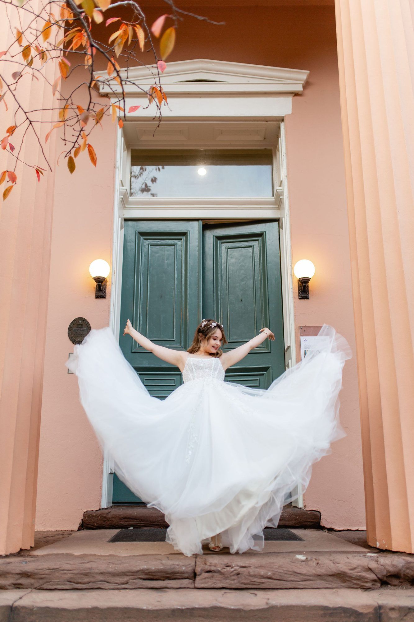 Bride in white tulle gown twirling before teal double doors of a pink building with autumn foliage.