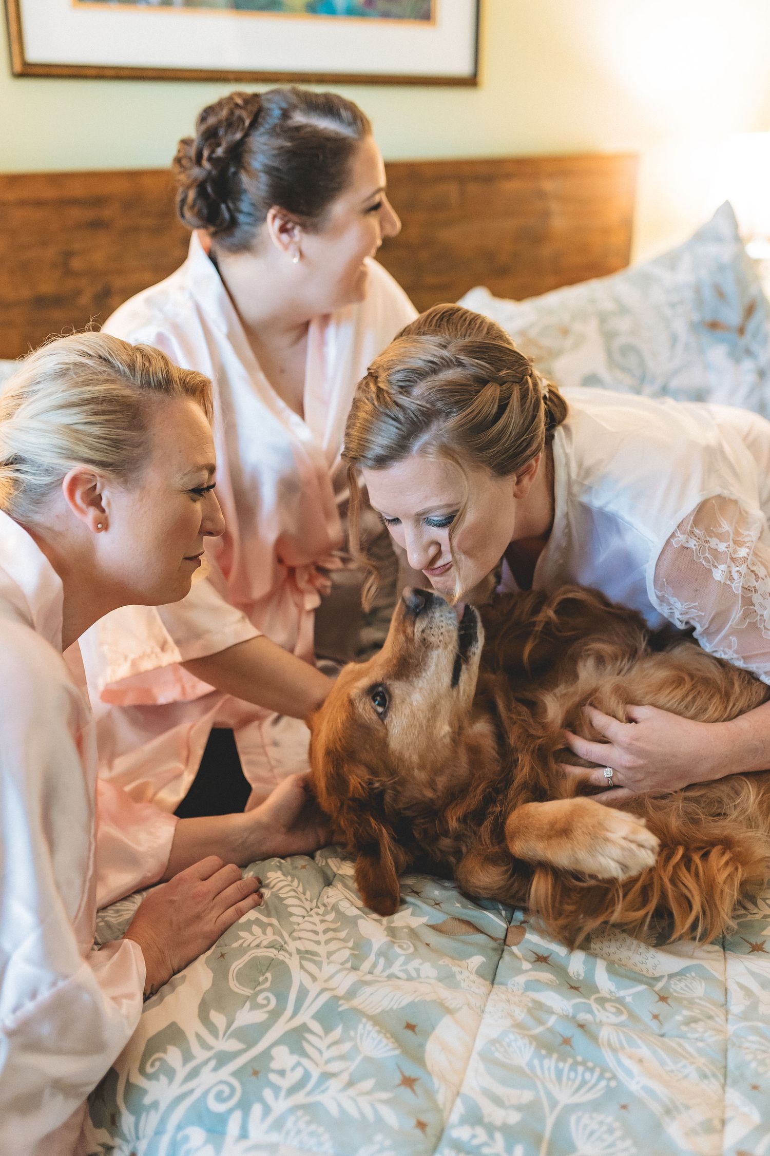 Three bridesmaids in robes cuddle with a golden cocker spaniel on a bed during wedding day preparations.