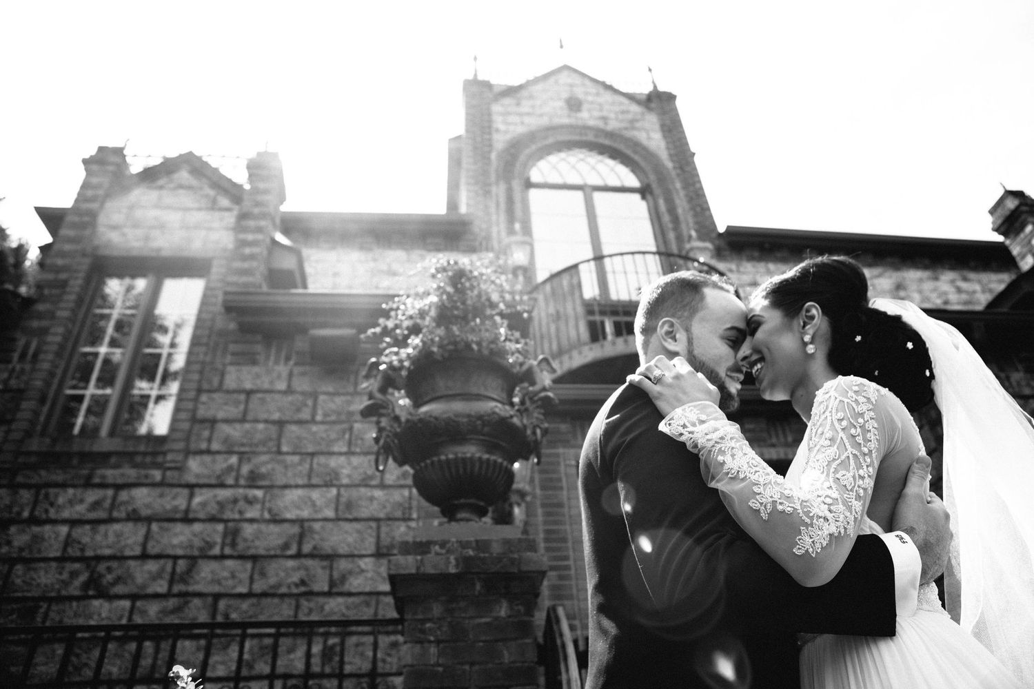 Bride and groom share a romantic embrace outside a stone mansion in a classic black and white wedding photo.