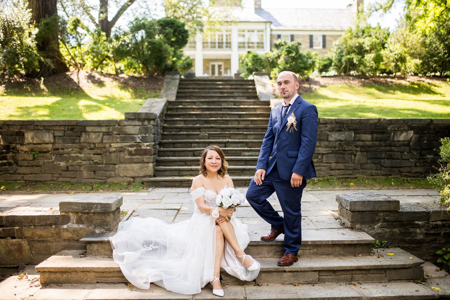 Bride in white gown sits on stone steps while groom in navy suit stands beside her at elegant estate wedding venue.