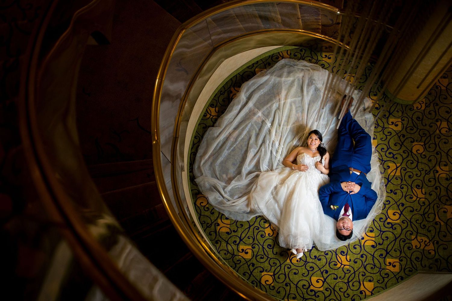 Aerial view of bride in white gown and groom in blue suit lying on ornate spiral staircase carpet at wedding venue.