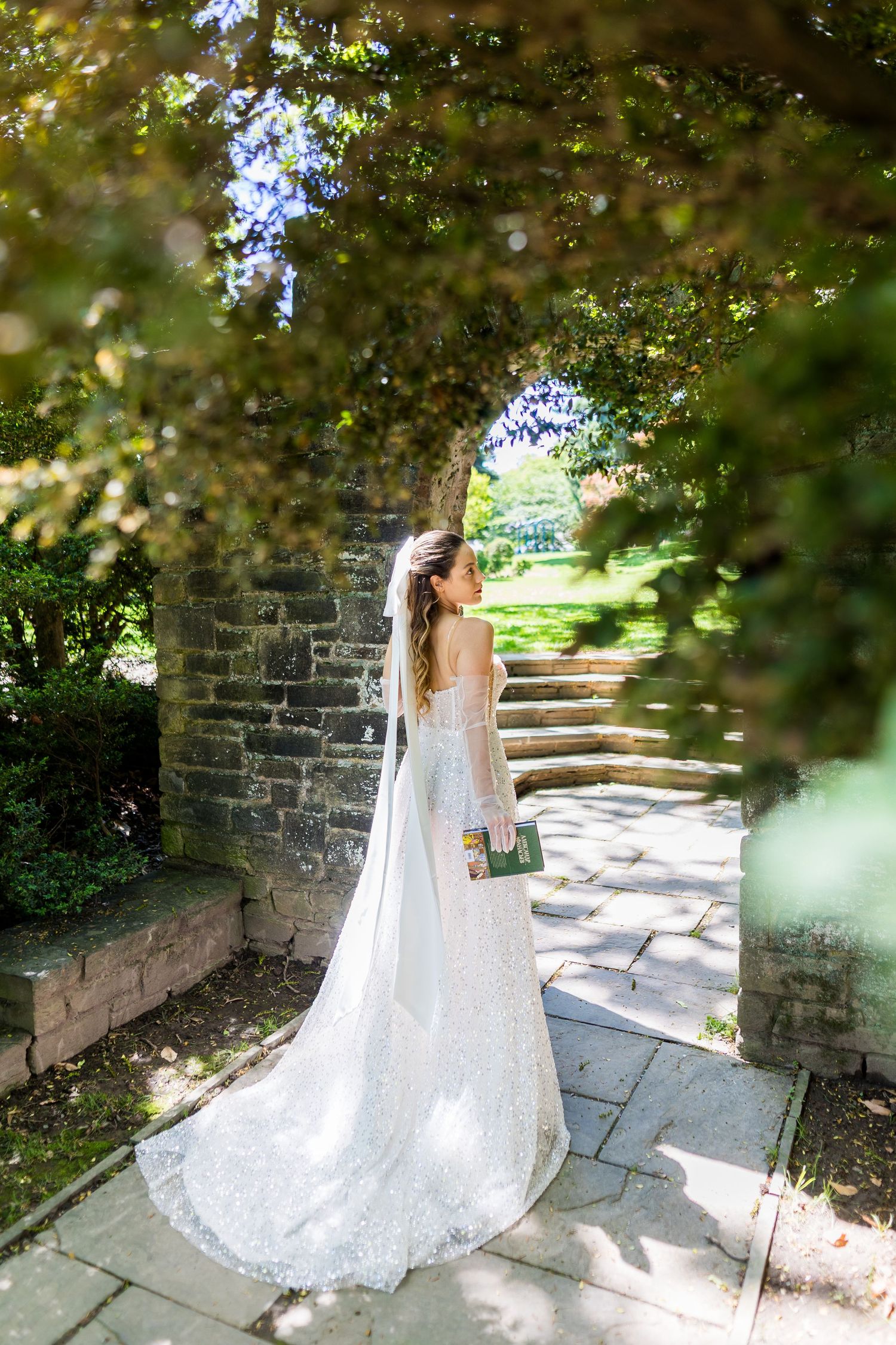 Bride in lace wedding gown with long veil stands under stone arch in a lush garden pathway.