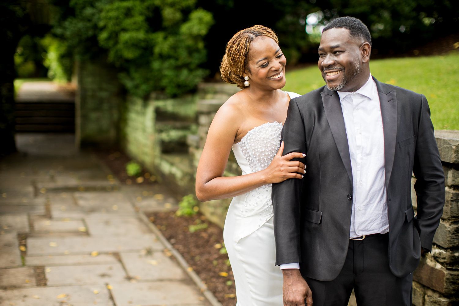 A joyful couple in wedding attire walks along a stone path, the bride in white and groom in a dark suit, smiling warmly.